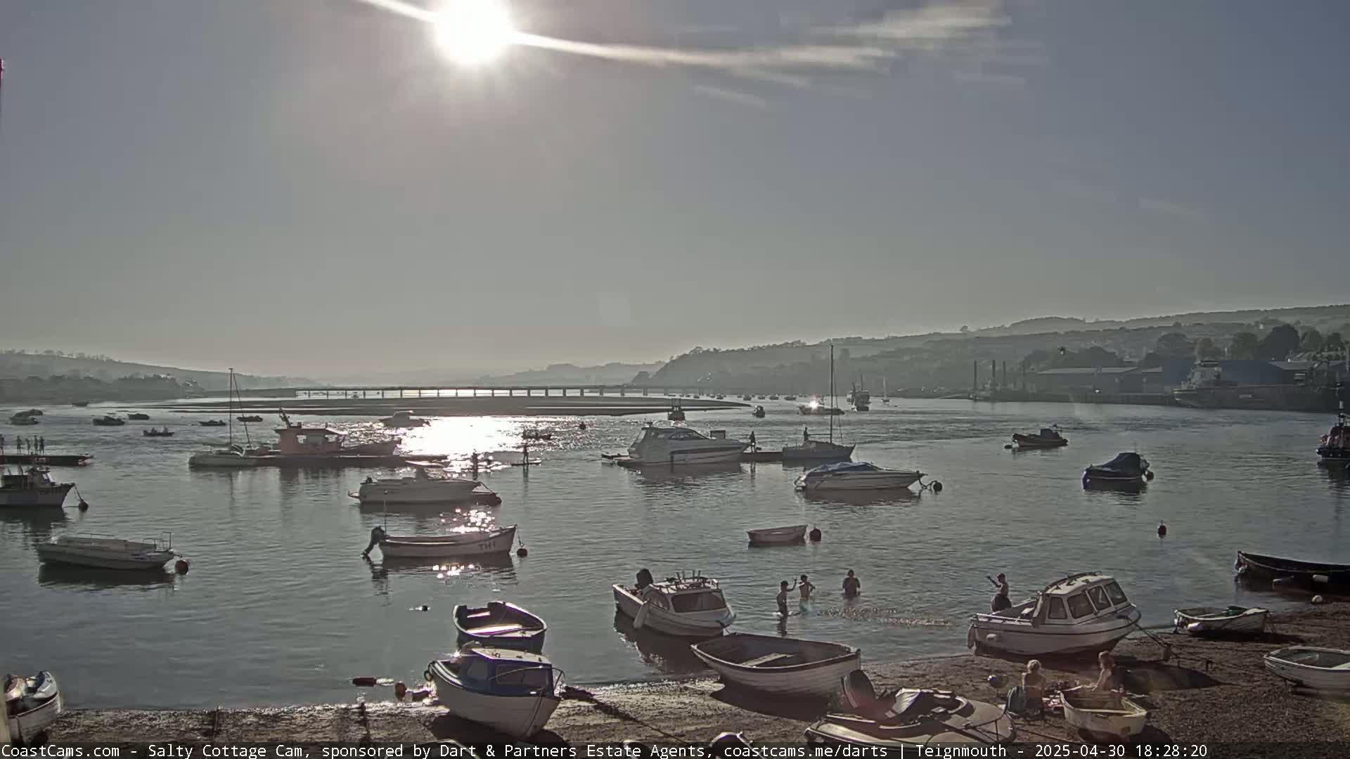 Numerous boats are moored in a calm, sunlit harbor, with several people swimming near a sandy shore.