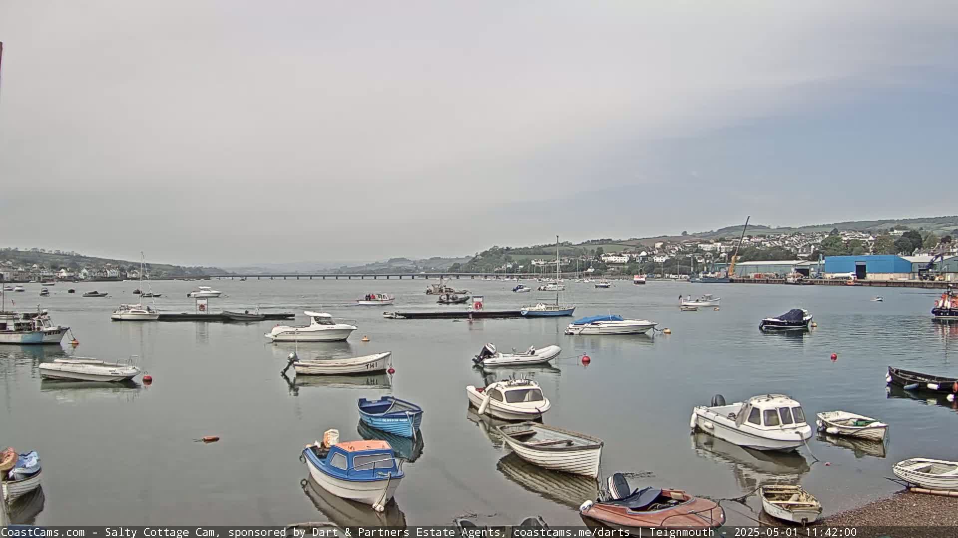 Many small motorboats are moored in a calm harbor under an overcast sky, with a bridge and distant town visible in the background.