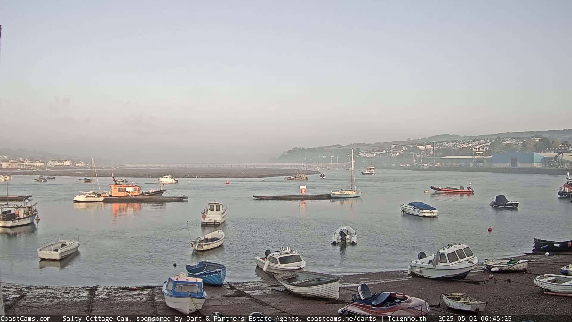 A calm, foggy morning reveals numerous small boats moored in a harbor, with a larger boat in the mid-ground and a distant shoreline visible.