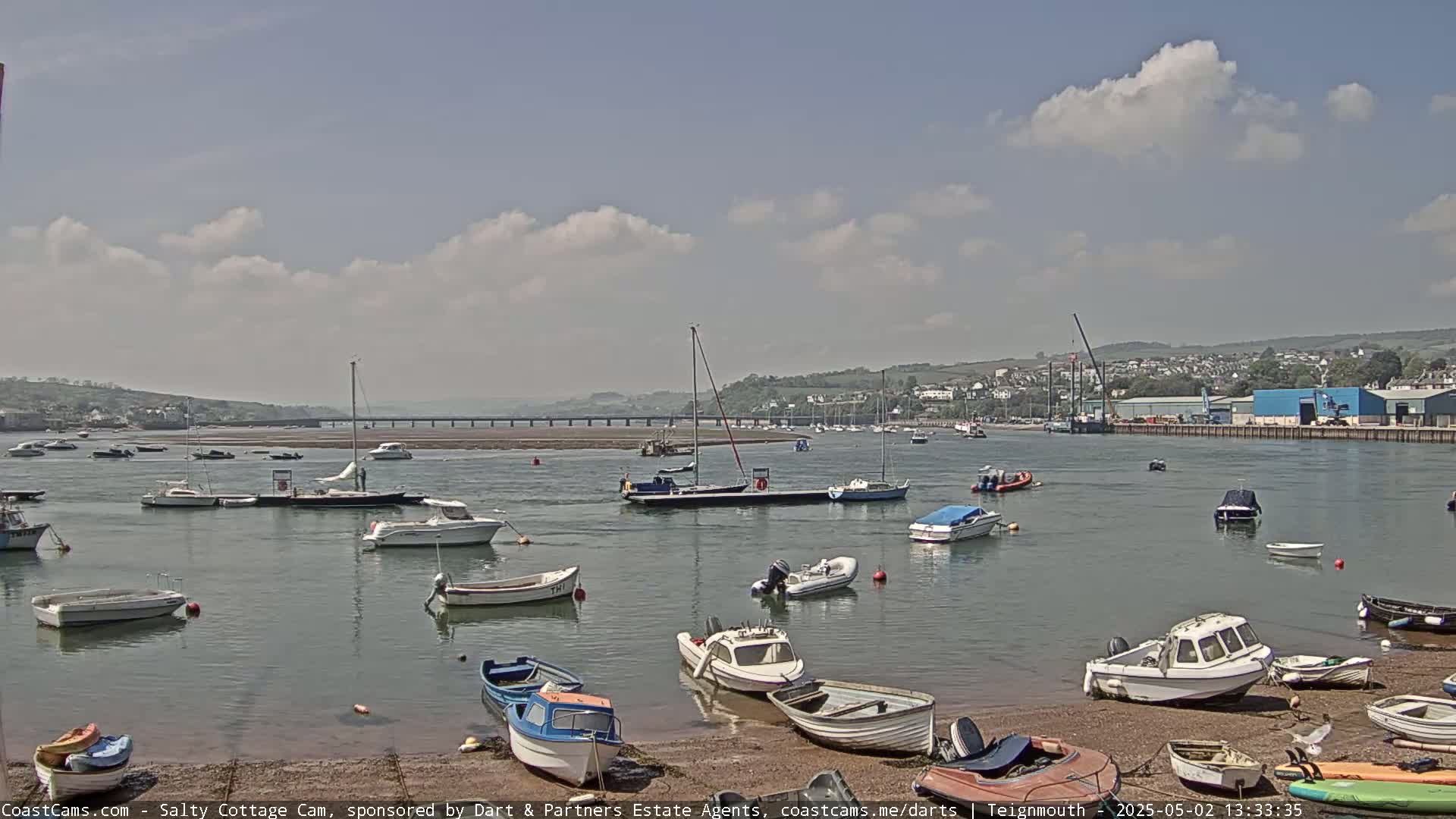 Numerous small boats are moored in calm water under a partly cloudy sky, with a bridge and buildings visible in the distance.