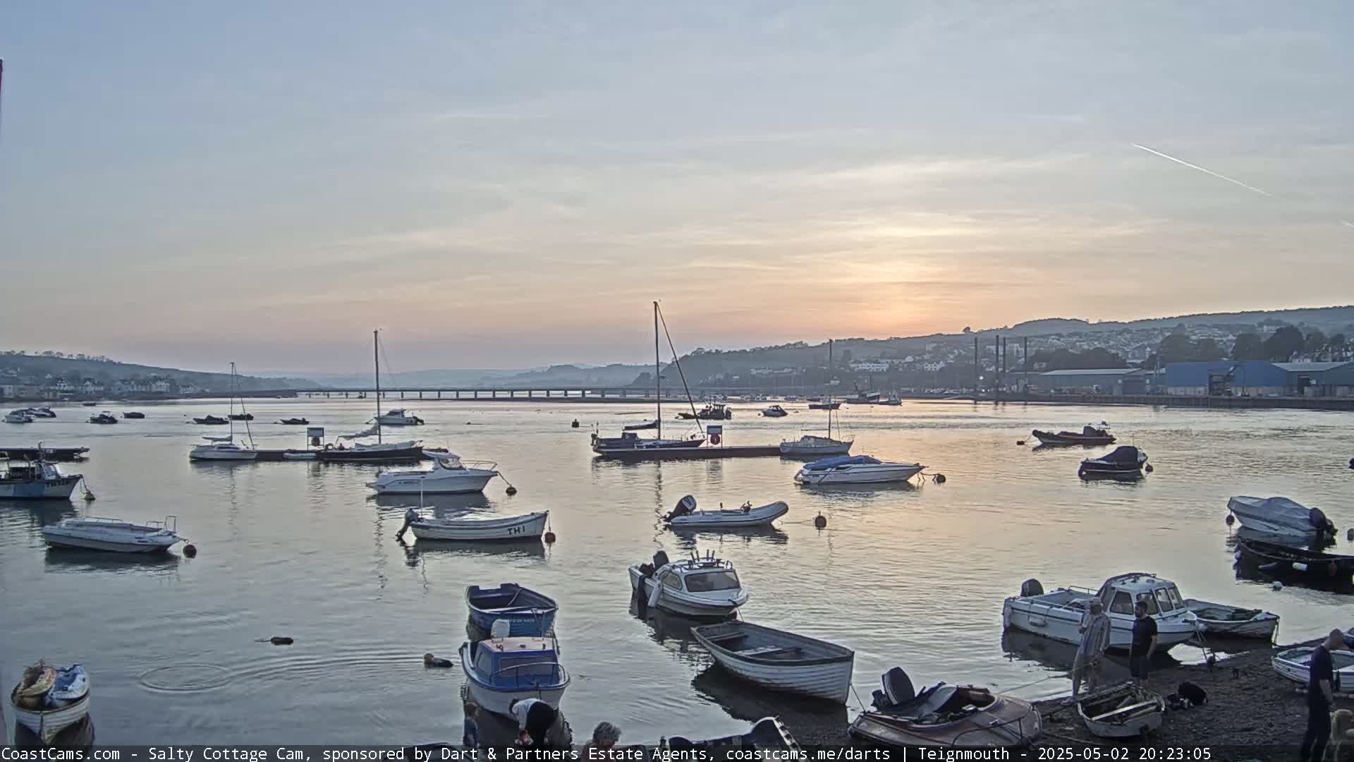Many small boats are moored in a calm harbor at sunset, with a town and bridge visible in the background.