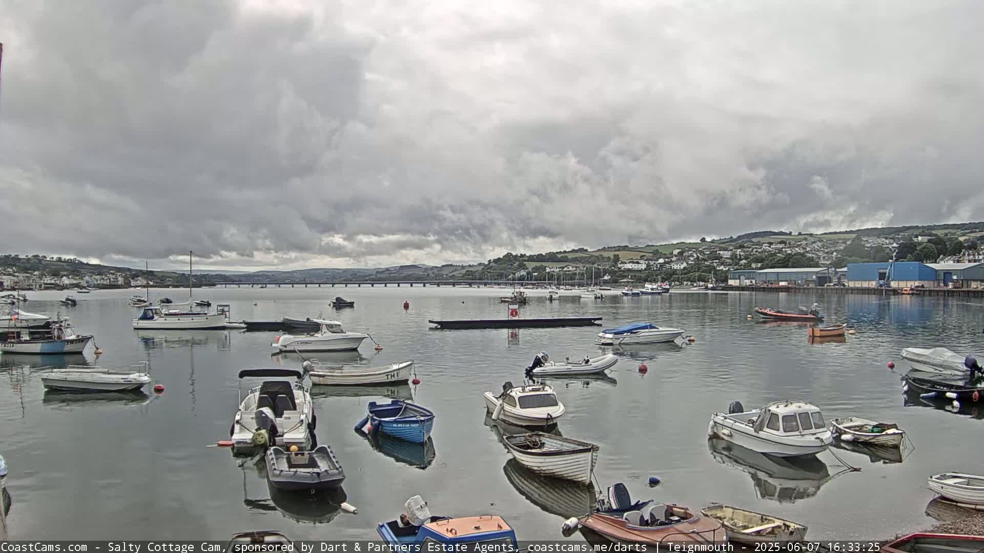 Numerous small boats are moored in calm water under a cloudy sky, with a bridge and distant hills visible in the background.