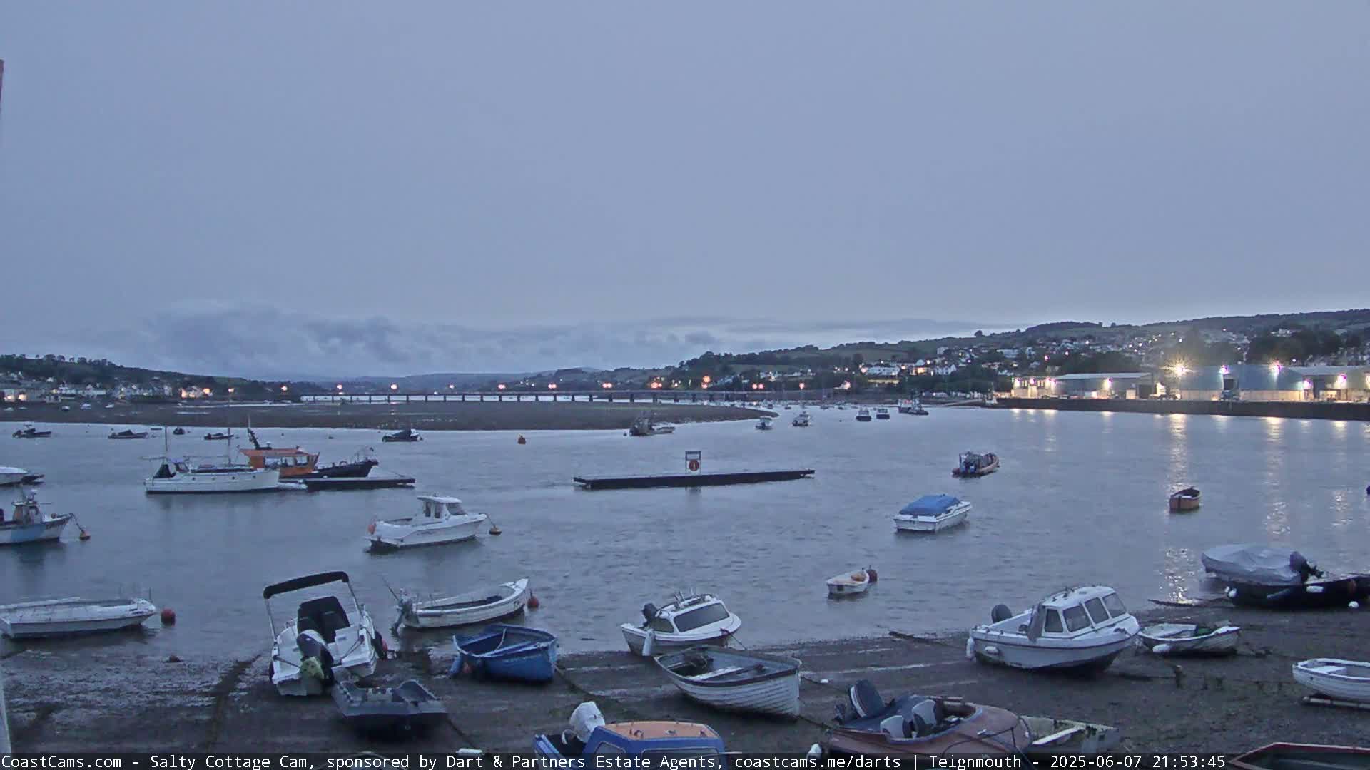 Numerous small boats are moored in a calm harbor at twilight, with a town and bridge visible in the background under a cloudy sky.