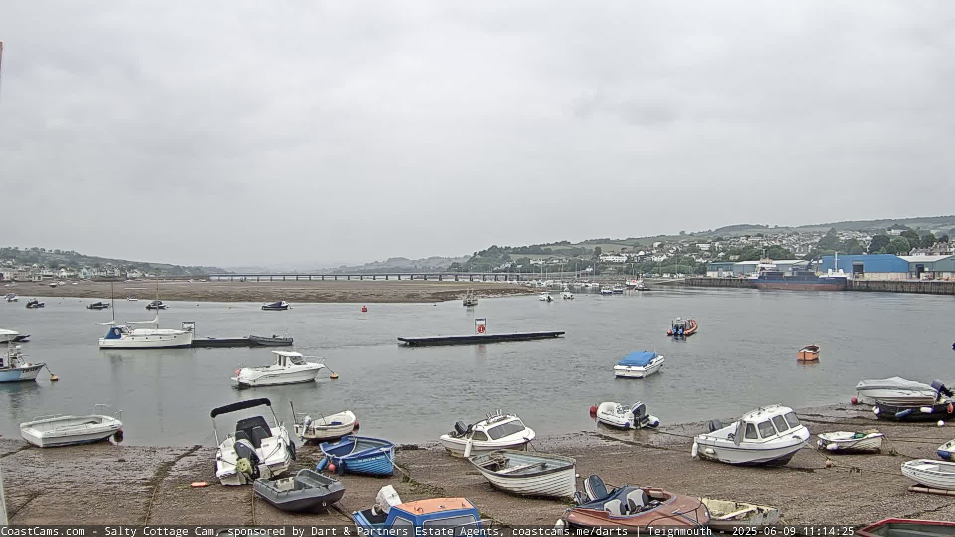Numerous small boats are moored along a shoreline and in the calm water of a harbor under an overcast sky, with a bridge and distant town visible in the background.
