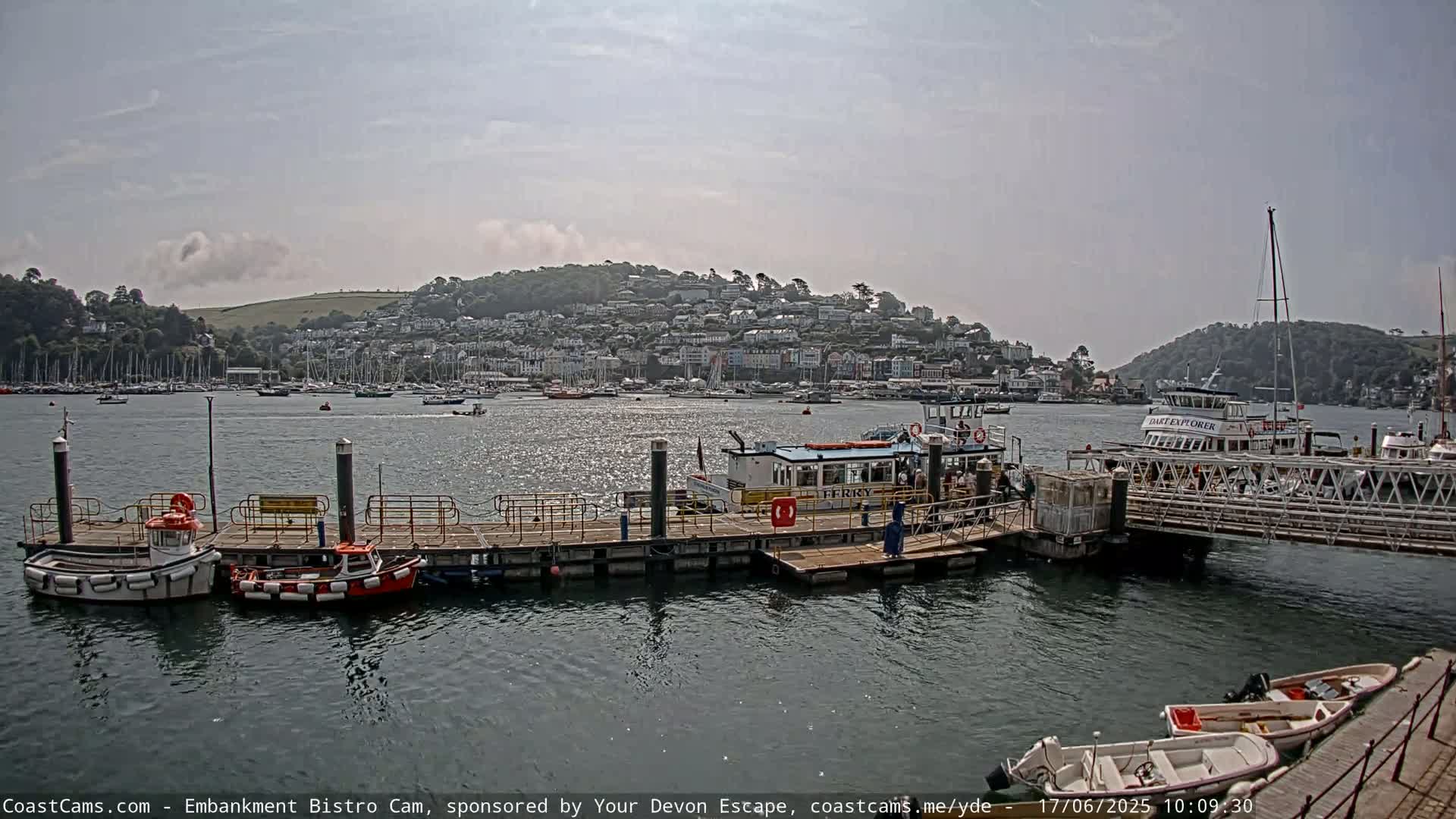 A ferry is docked at a pier in a harbor, with numerous other boats and a town nestled on a hillside in the background under a partly cloudy sky.