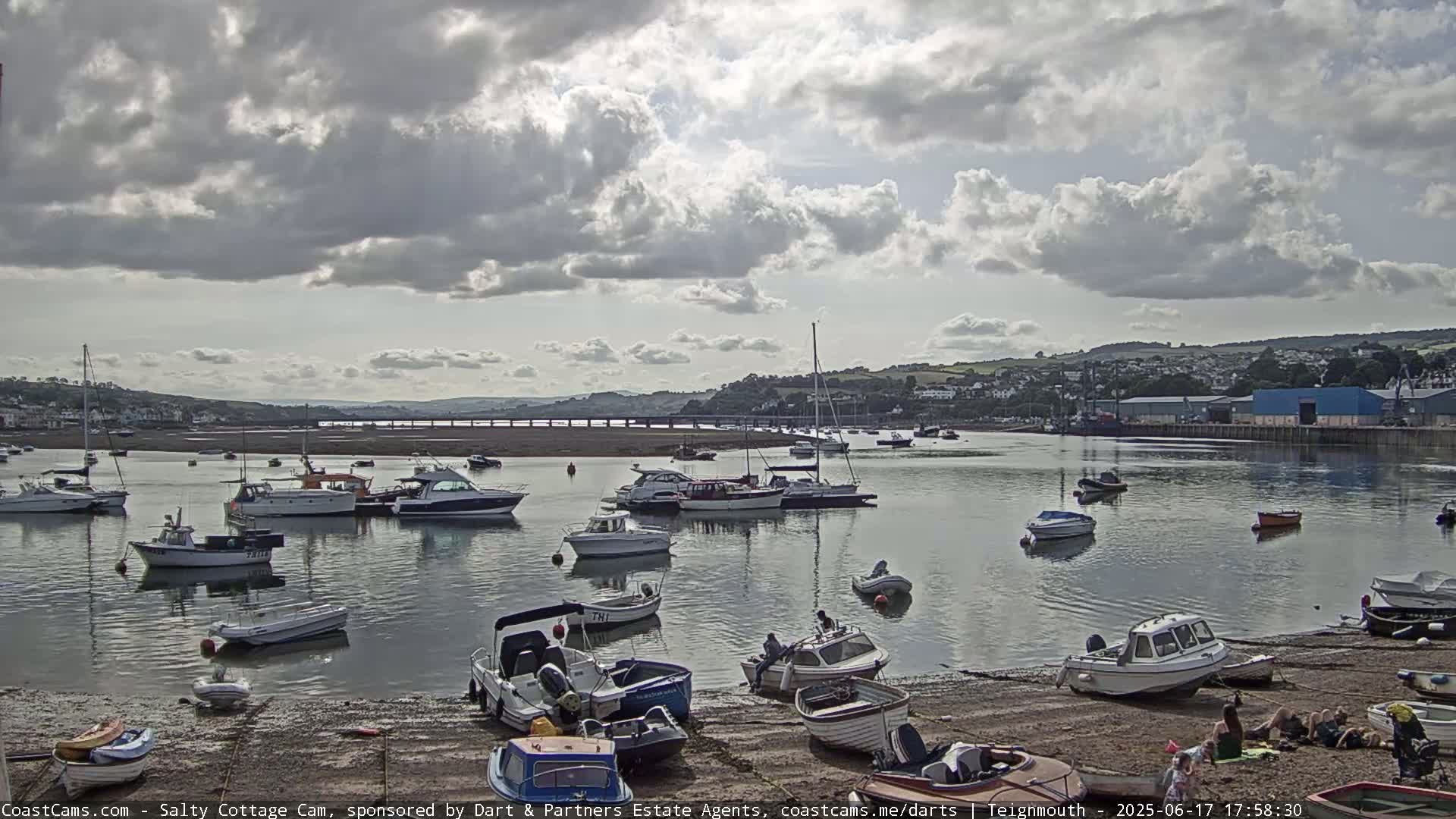 Numerous boats are moored in a calm harbor under a partly cloudy sky, with a shoreline visible in the foreground.