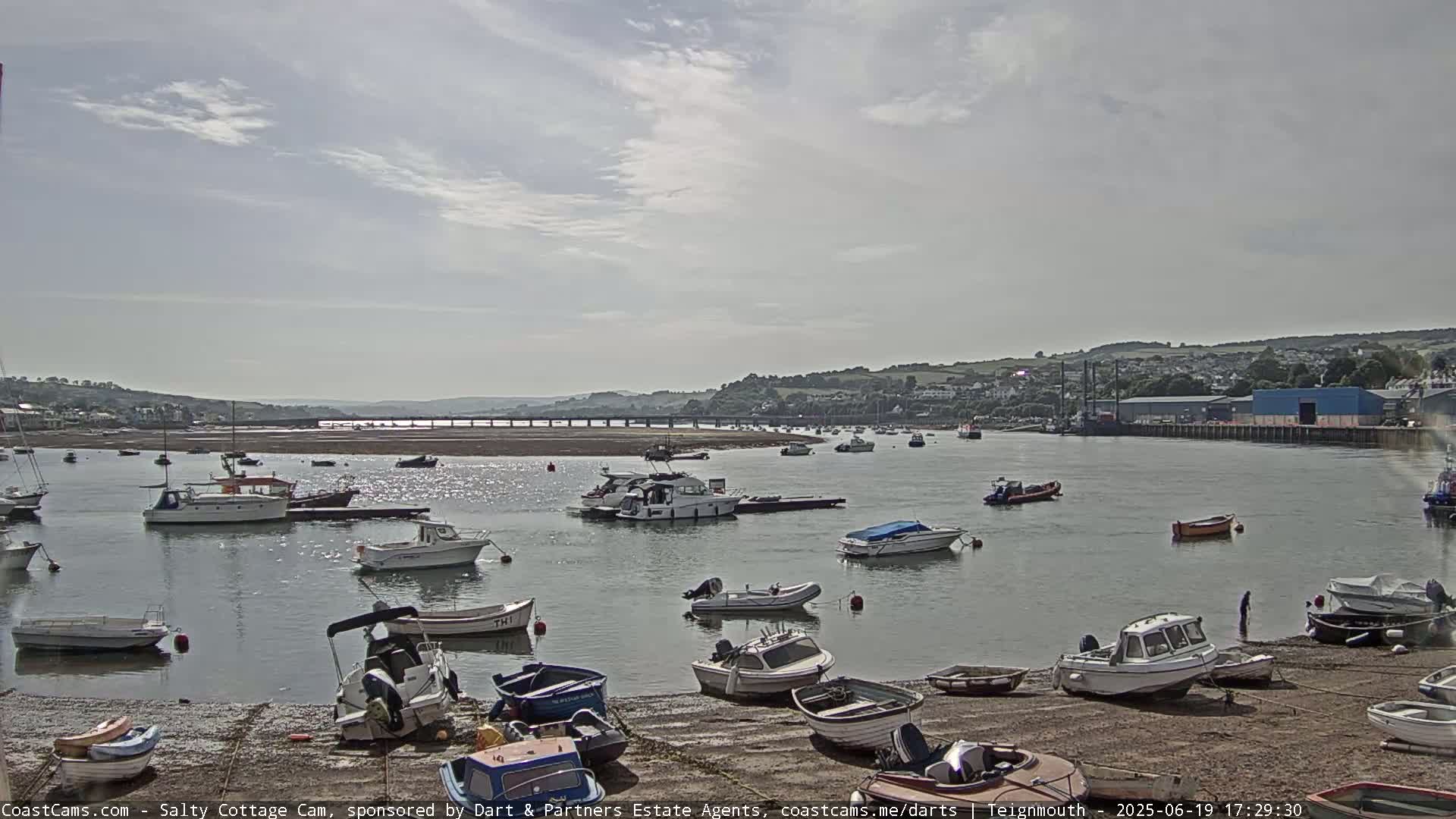 A mostly sunny day shows numerous boats in a calm harbor, some on the shore and others on the water, with a bridge and distant hills visible in the background.