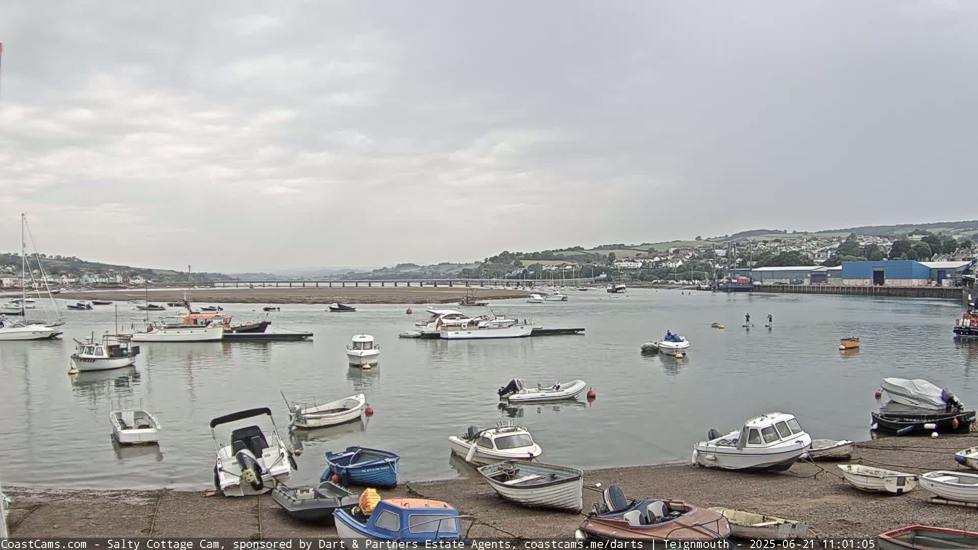 Many small motorboats and sailboats are moored in a calm harbor on an overcast day, with a town and bridge visible in the distance.