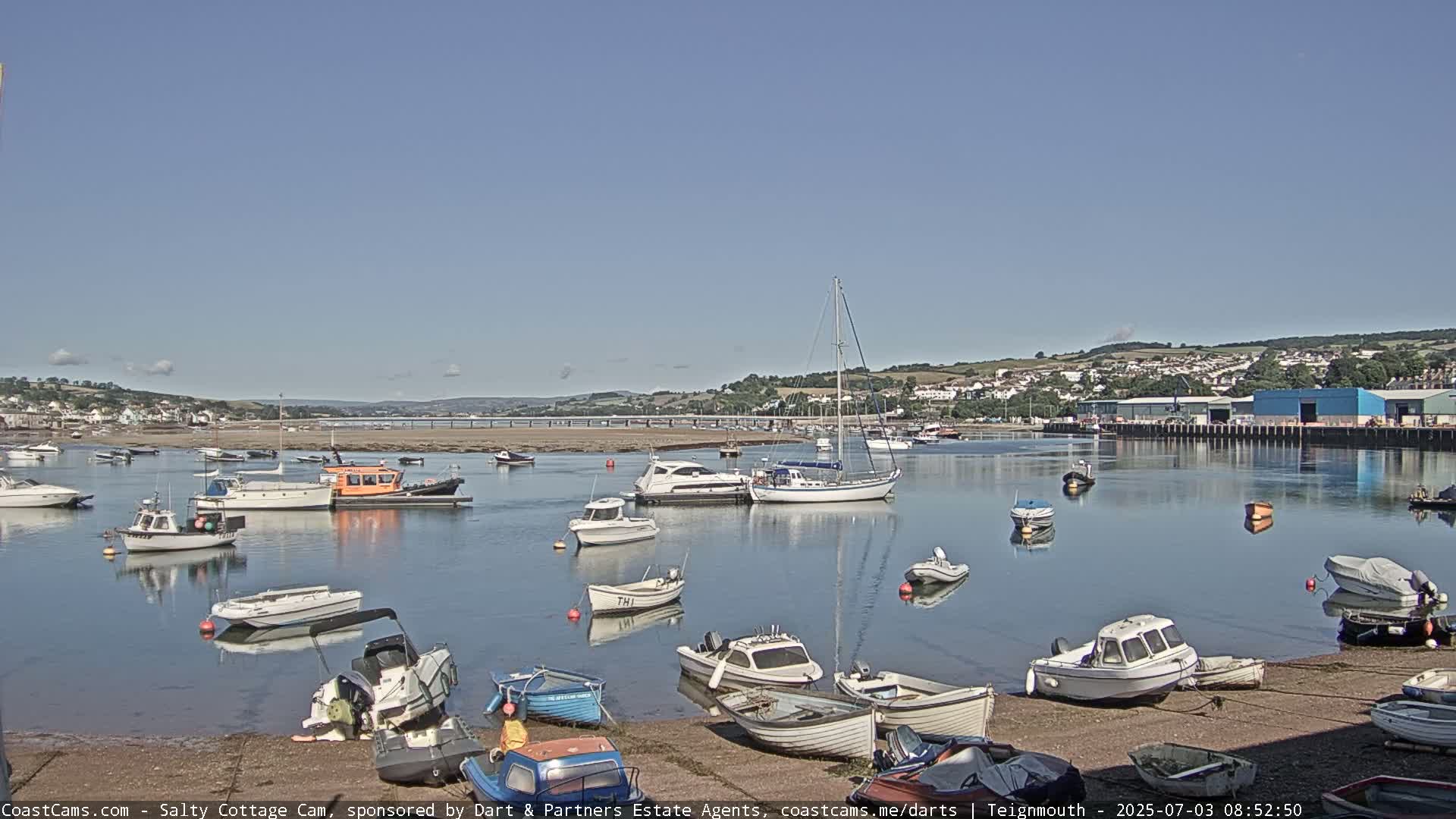 A calm harbor with numerous small boats and yachts is nestled against a shoreline under a clear, sunny sky.