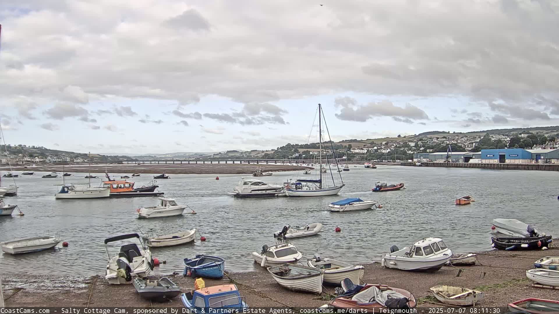 Numerous small boats are moored in a calm harbor under a cloudy sky, with a larger sailboat and a few other vessels further out in the water.