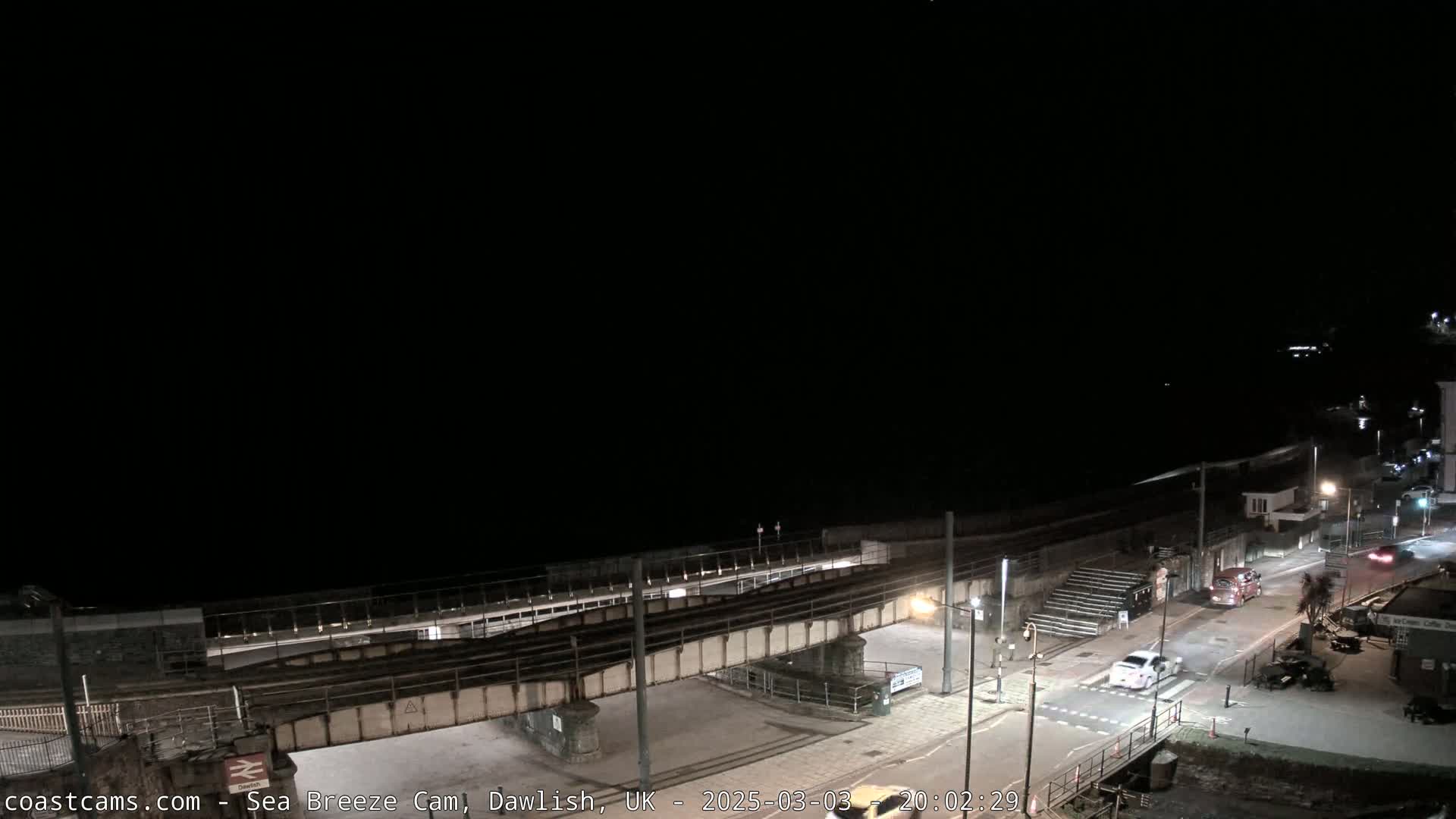 A nighttime view of a coastal town with a railway bridge over a road leading to the ocean under a dark sky.