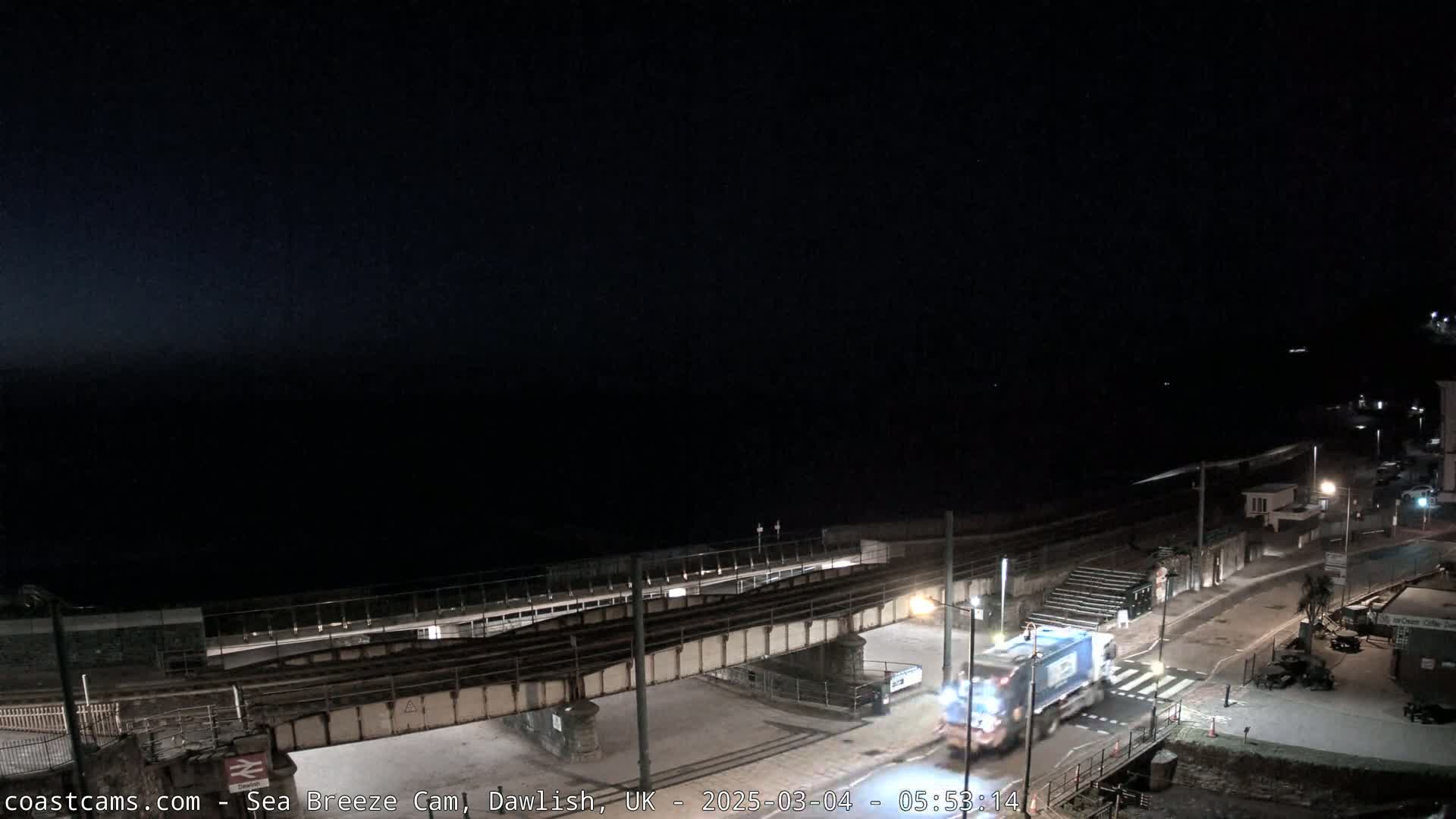 A nighttime coastal scene shows a garbage truck driving on a road that passes under a railway bridge, with a dark ocean in the background under a clear, starless sky.