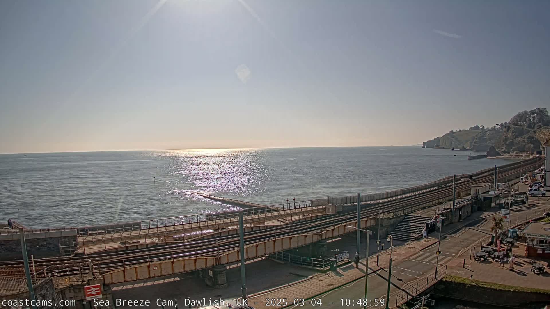 A sunny day shows a coastal scene with a railway line running along a sea wall next to a road and buildings, overlooking a calm, sunlit sea.