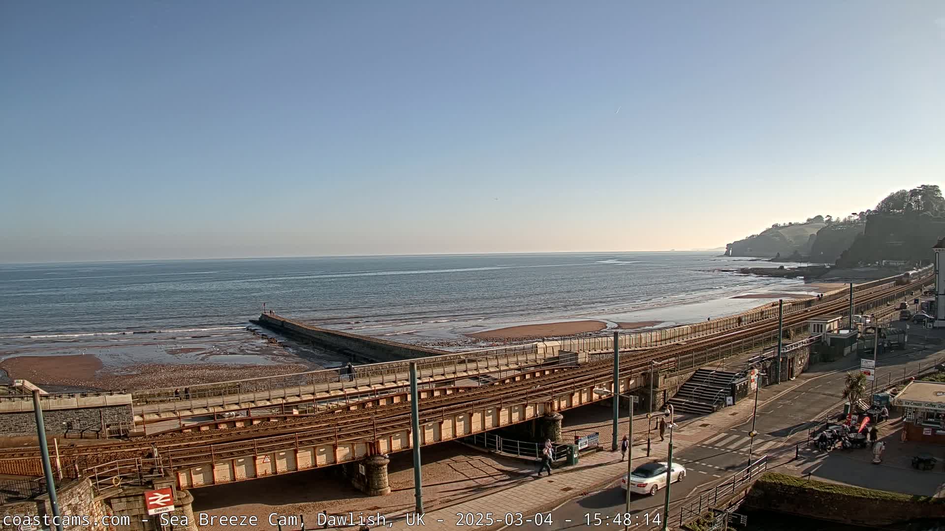 A railway line runs along a sandy beach next to a calm ocean under a clear blue sky.