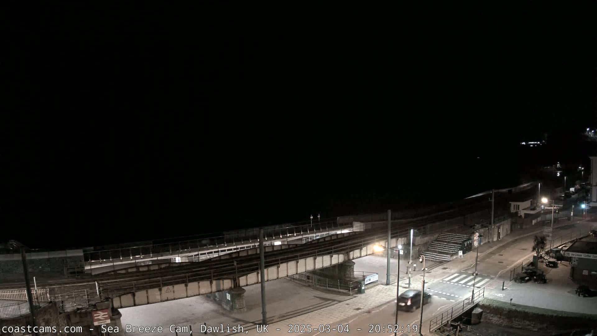 A nighttime, overhead view of a coastal town's train tracks and road intersecting at a pedestrian crossing, with a dark ocean in the background under a clear, black sky.