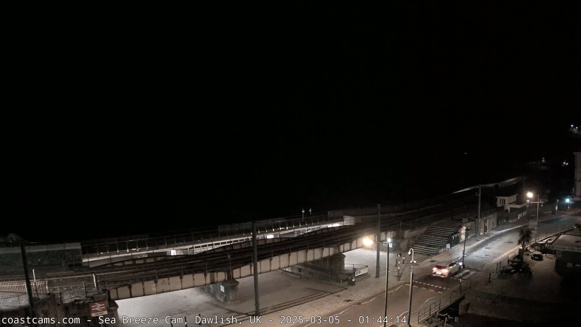 A nighttime coastal scene shows a railway bridge over a road next to a dark beach under a clear, dark sky.