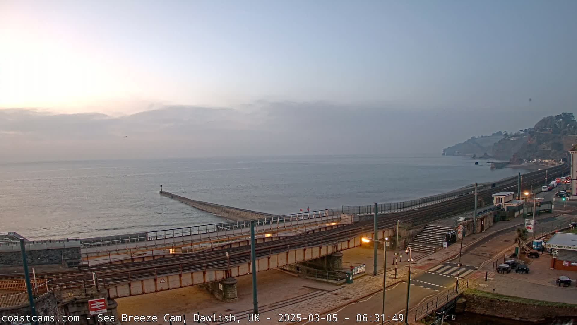 A hazy sunrise over a calm sea, viewed from above a coastal railway line and road leading to a seawall and cliffs.