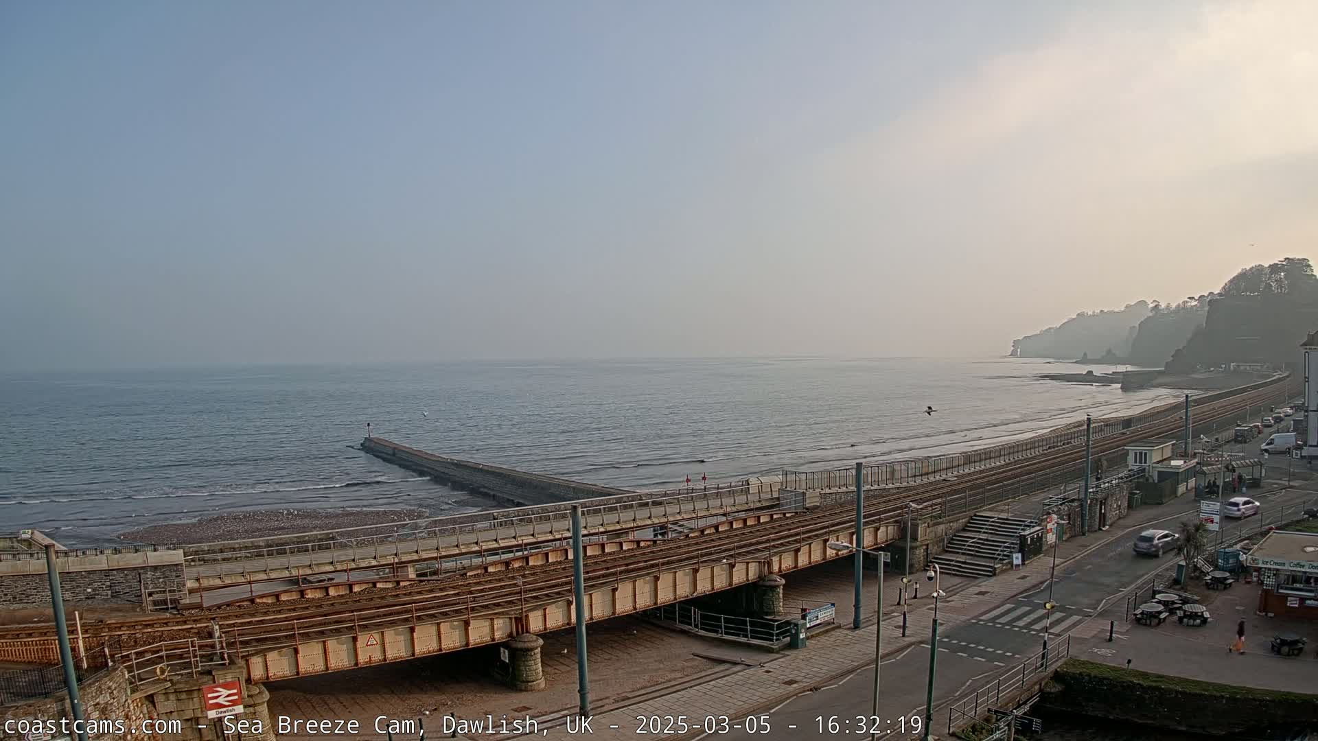 A hazy, overcast day reveals a coastal scene with train tracks crossing a beach and leading to a cliffside, a breakwater extending into the sea.