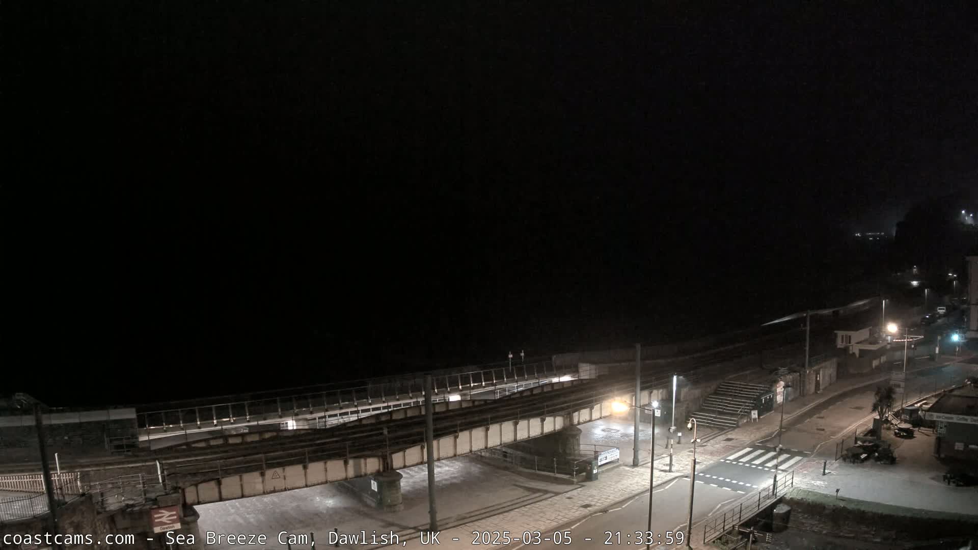 A nighttime, dark view of a coastal town with a railway bridge over a road and a mostly-dark sea in the background.