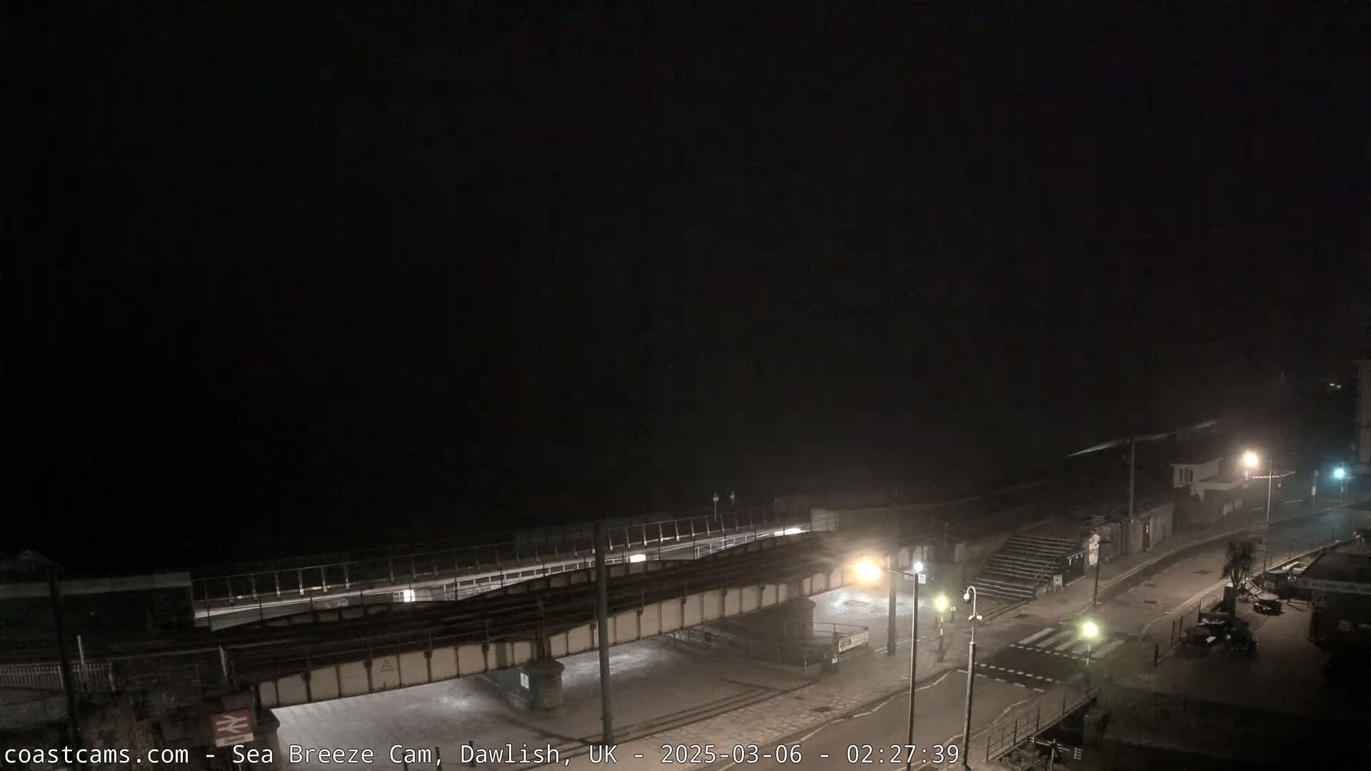 A nighttime coastal scene shows a railway bridge over a road leading to the sea, under dark, foggy conditions.