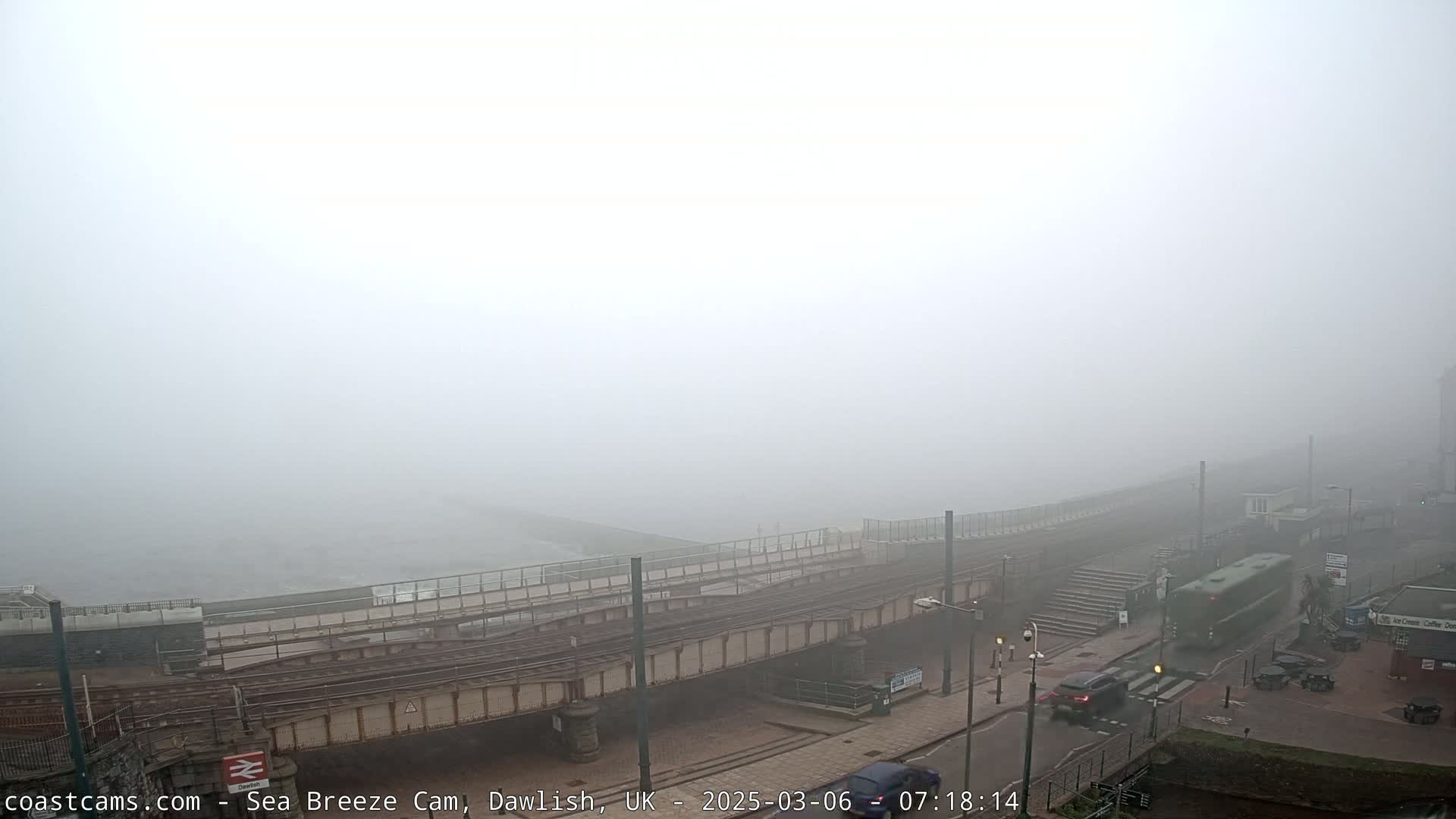 A thick fog obscures the view of a coastal railway line and a street, with a few vehicles visible near buildings.