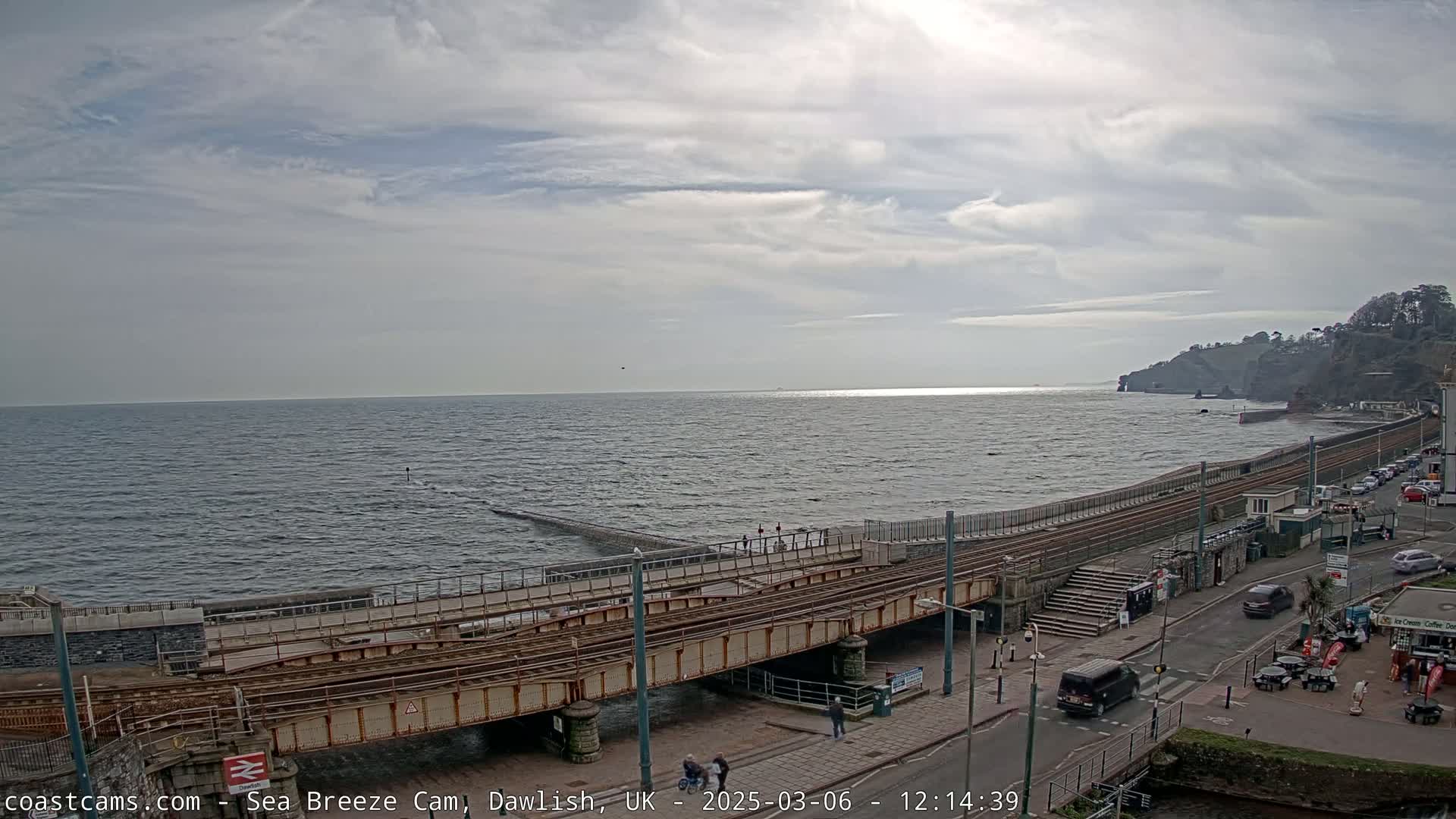 A partly cloudy day shows a seaside town with train tracks crossing a bridge over a rocky shoreline and calm ocean.