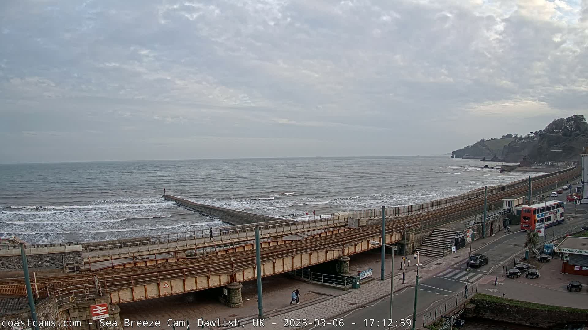 A coastal scene shows a railway track running alongside a beach with moderate waves under an overcast sky.