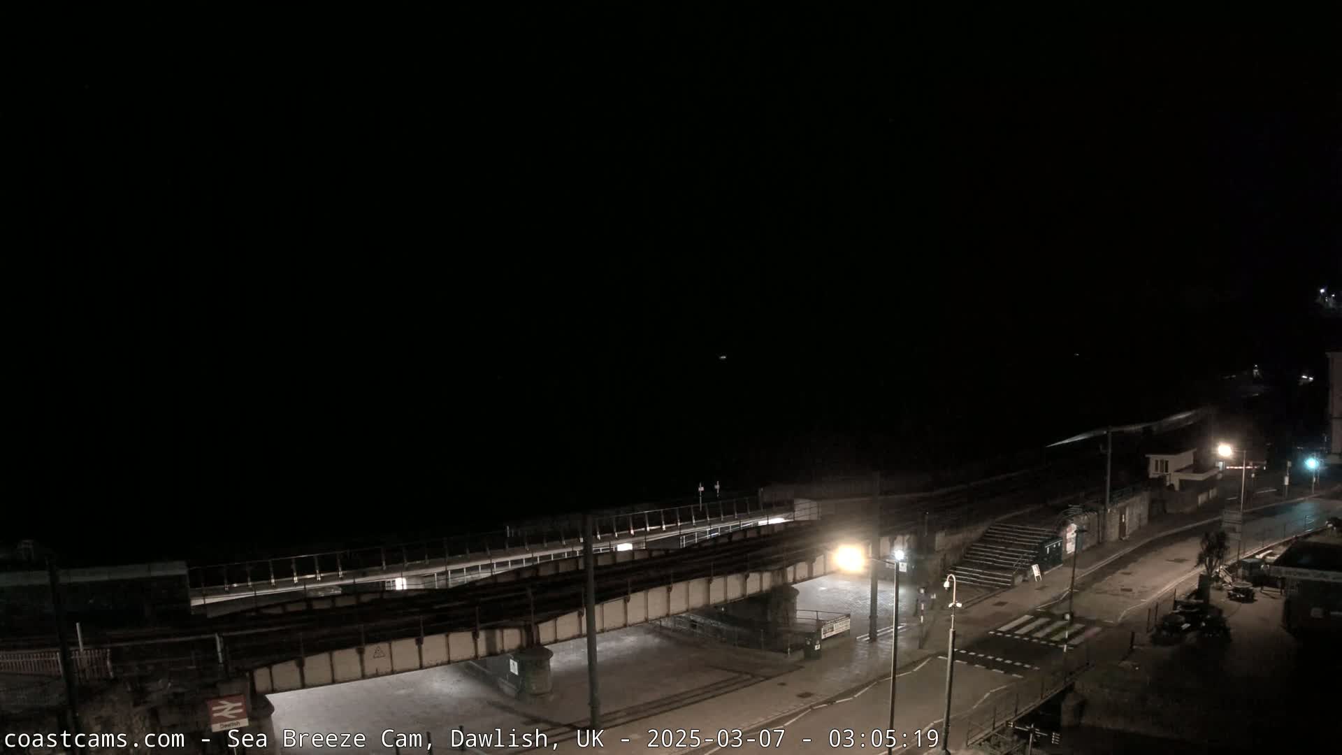 A nighttime, dark view of a coastal town, showing a railway bridge over a road leading to a beach under a clear, dark sky.