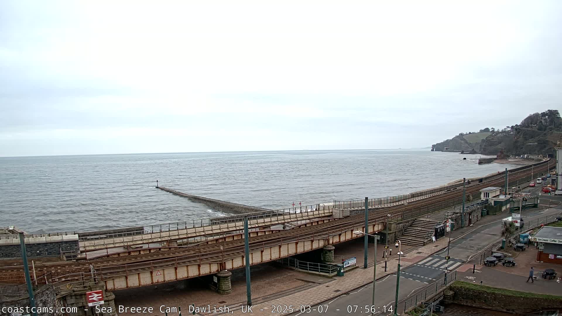 A coastal scene on an overcast day shows a railway track extending along a sea wall, with a breakwater jutting into a choppy sea.