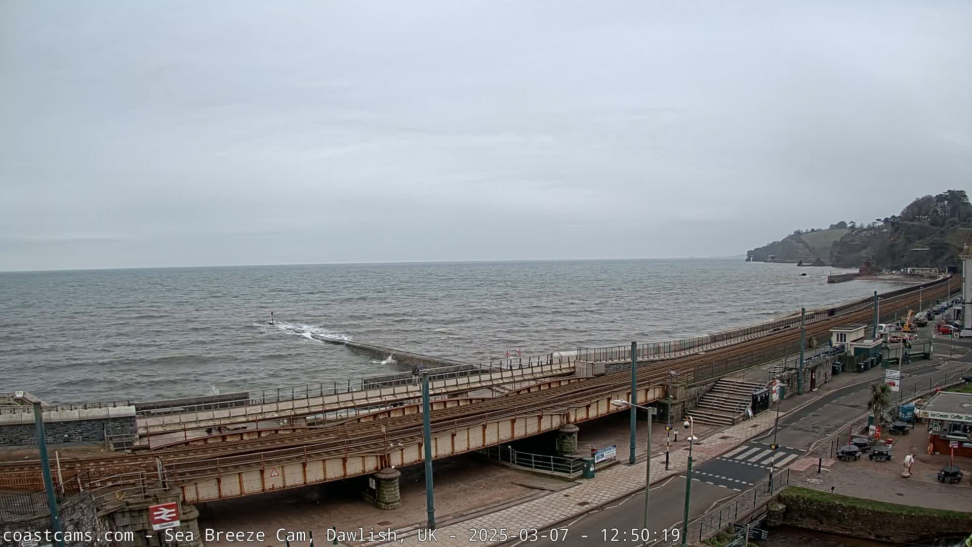 A choppy sea meets a coastal town with a railway line running along the shore under an overcast sky.