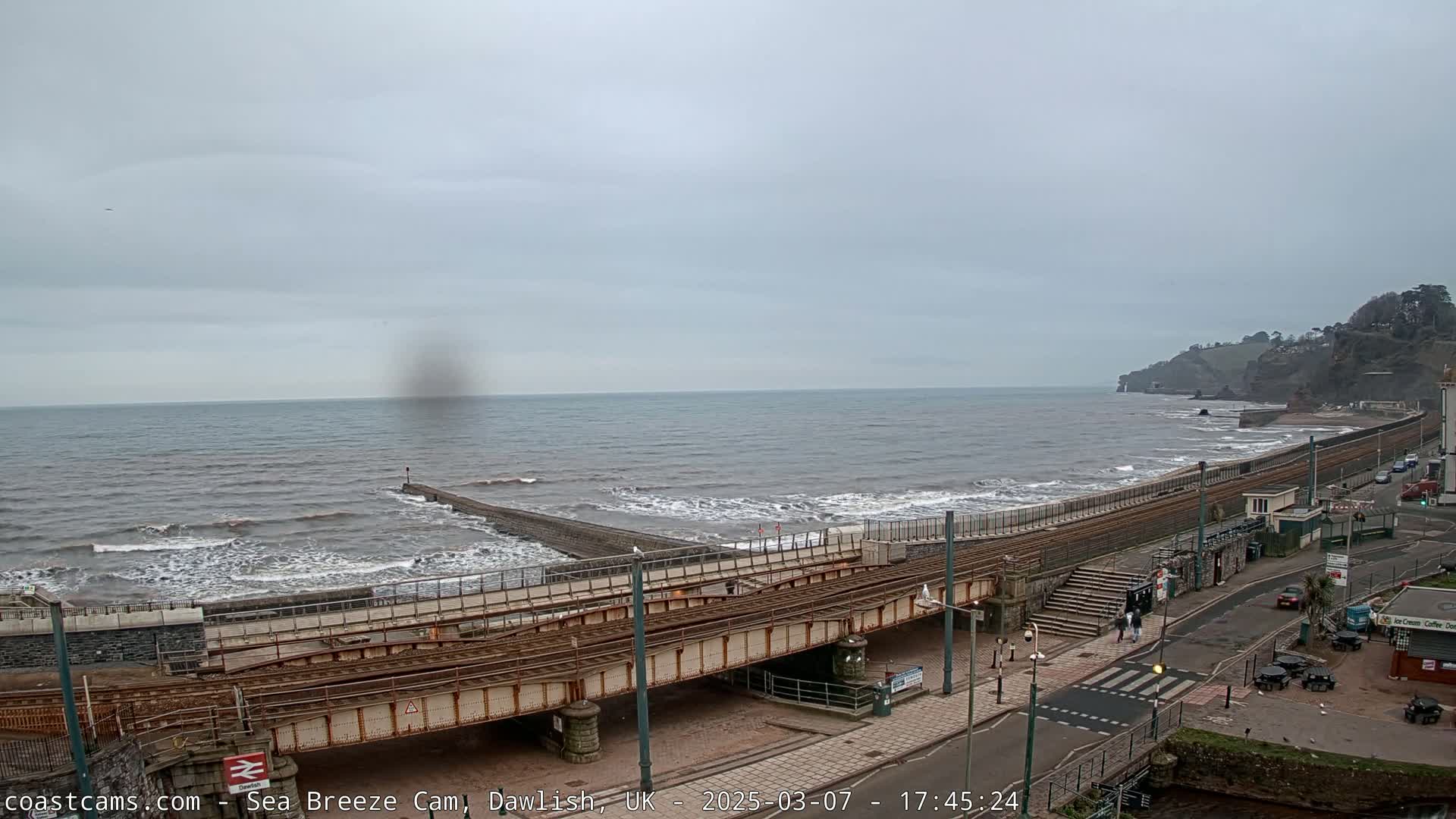 A seaside town with a railway line running along the coast, under an overcast sky and moderate waves crashing on the shore.
