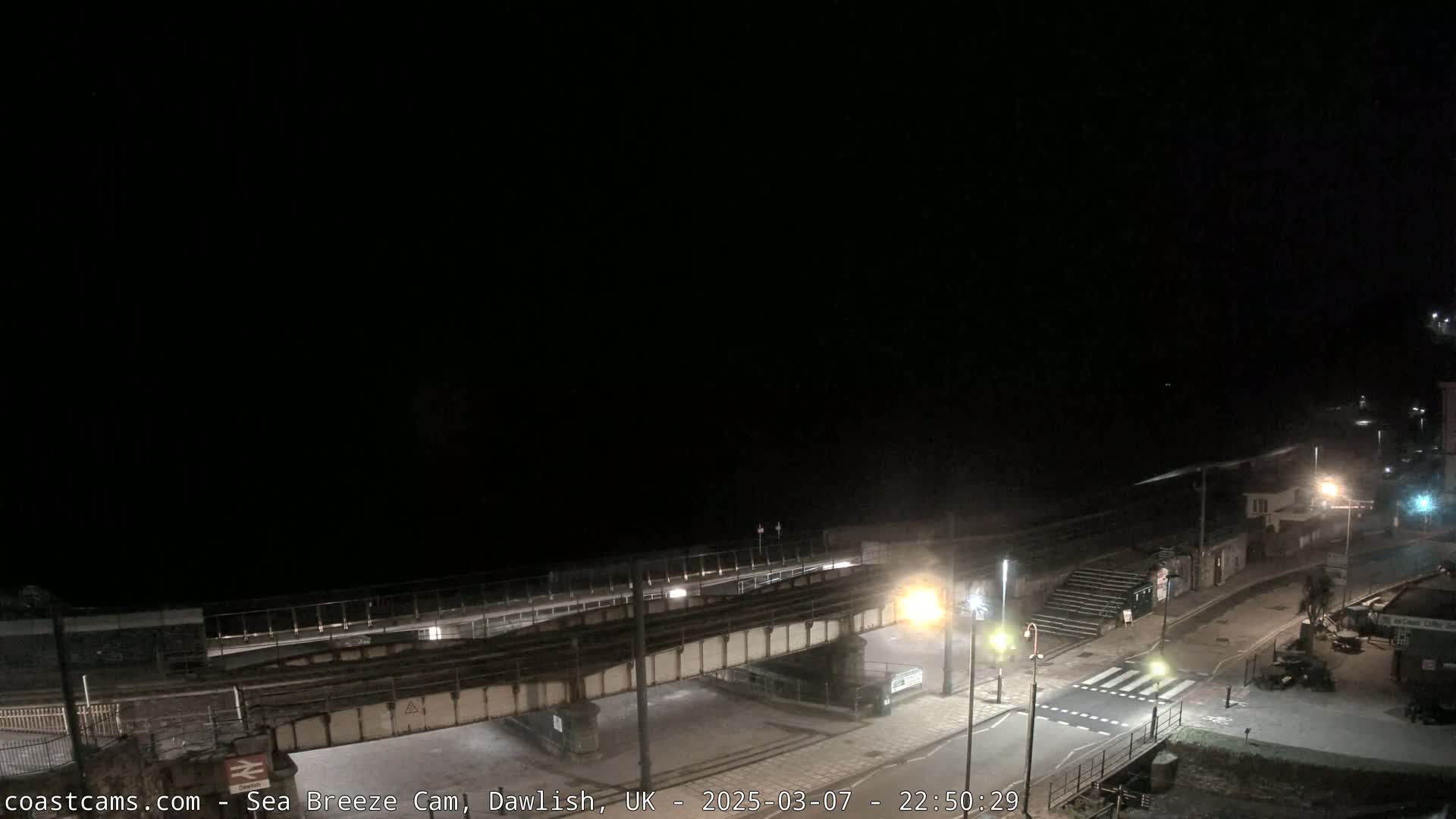 A nighttime, coastal view shows a railway bridge over a road and pedestrian crossing leading to a dark beach under a clear, black sky.