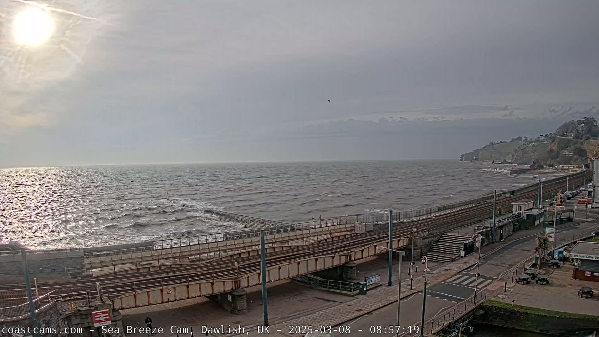 A mostly sunny day shows a choppy sea meeting a shoreline with train tracks and a coastal road.