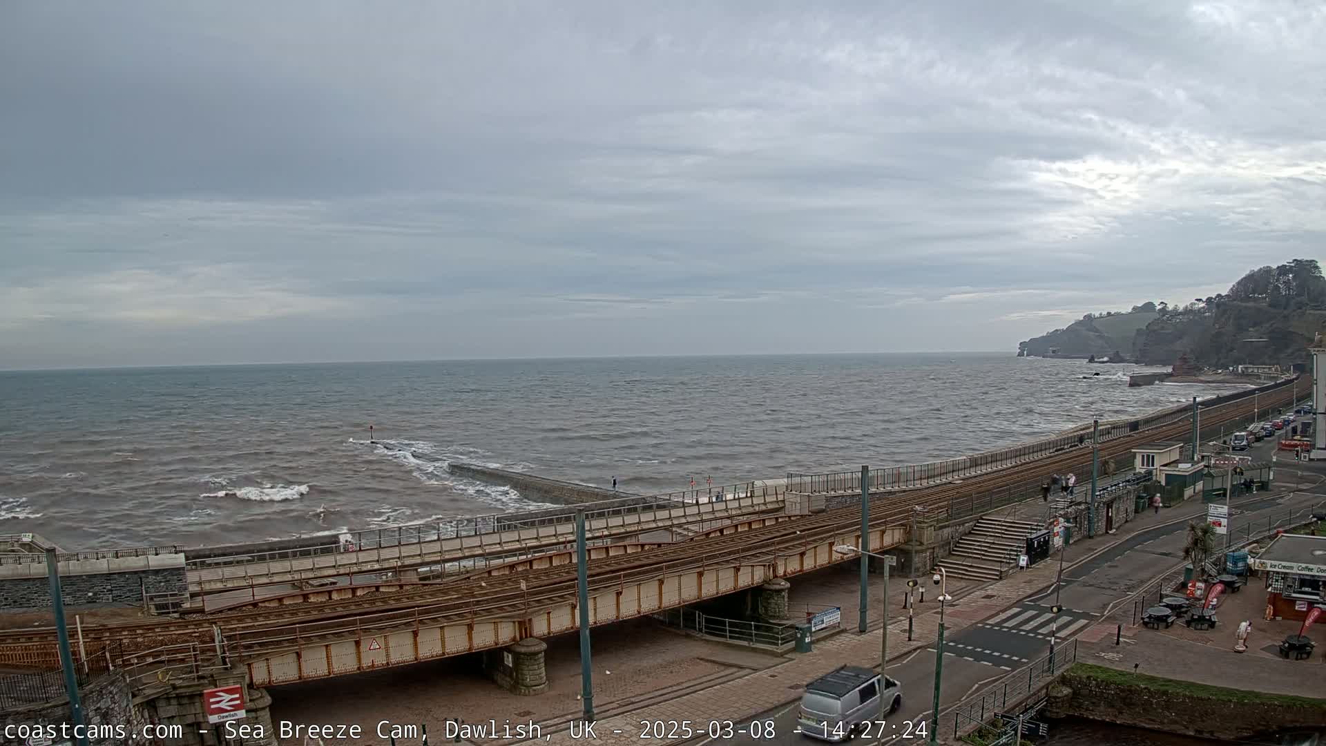 A railway line crosses a bridge over a road next to a sea wall, with a choppy sea under an overcast sky.