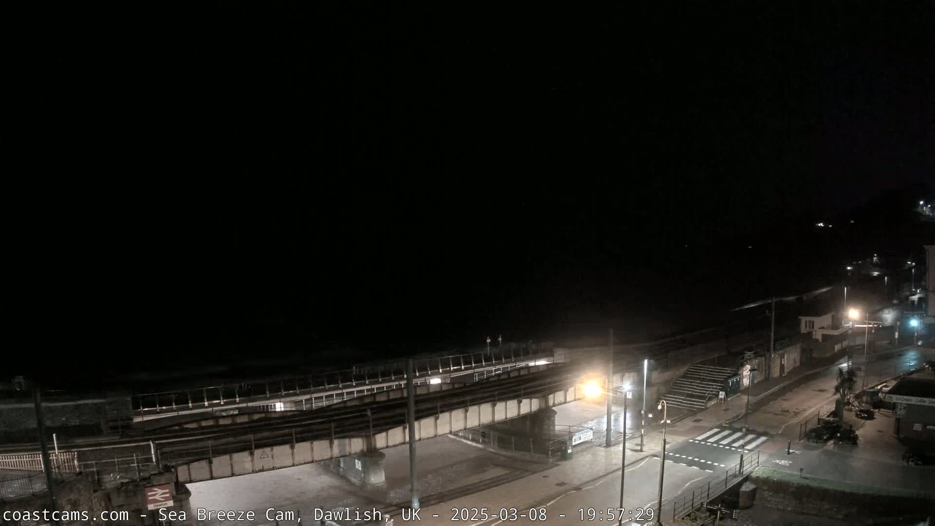 A nighttime view of a mostly empty seaside promenade and beach, with a bridge crossing over the road and railway line.