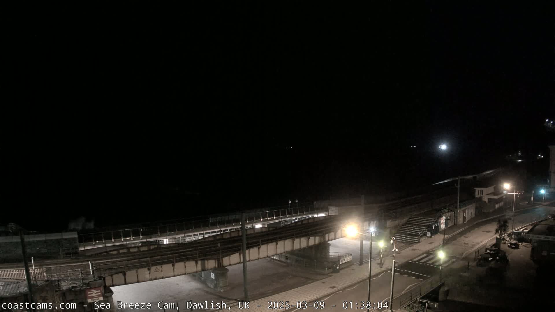 A nighttime, coastal scene shows a railway bridge crossing a road leading to a dark beach under a clear sky.