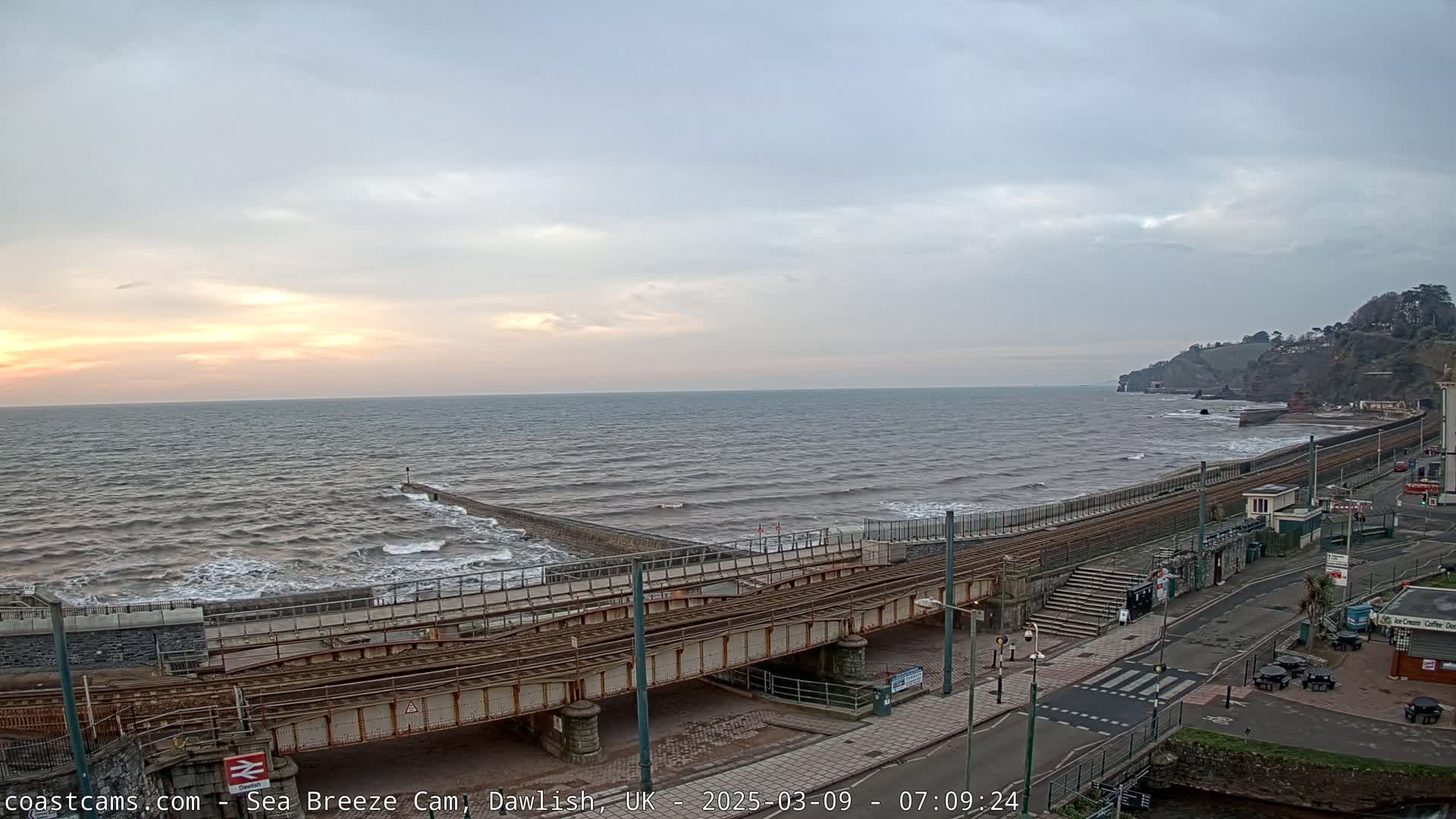 A partly cloudy sunset over a wavy sea next to a coastal railway line and road.