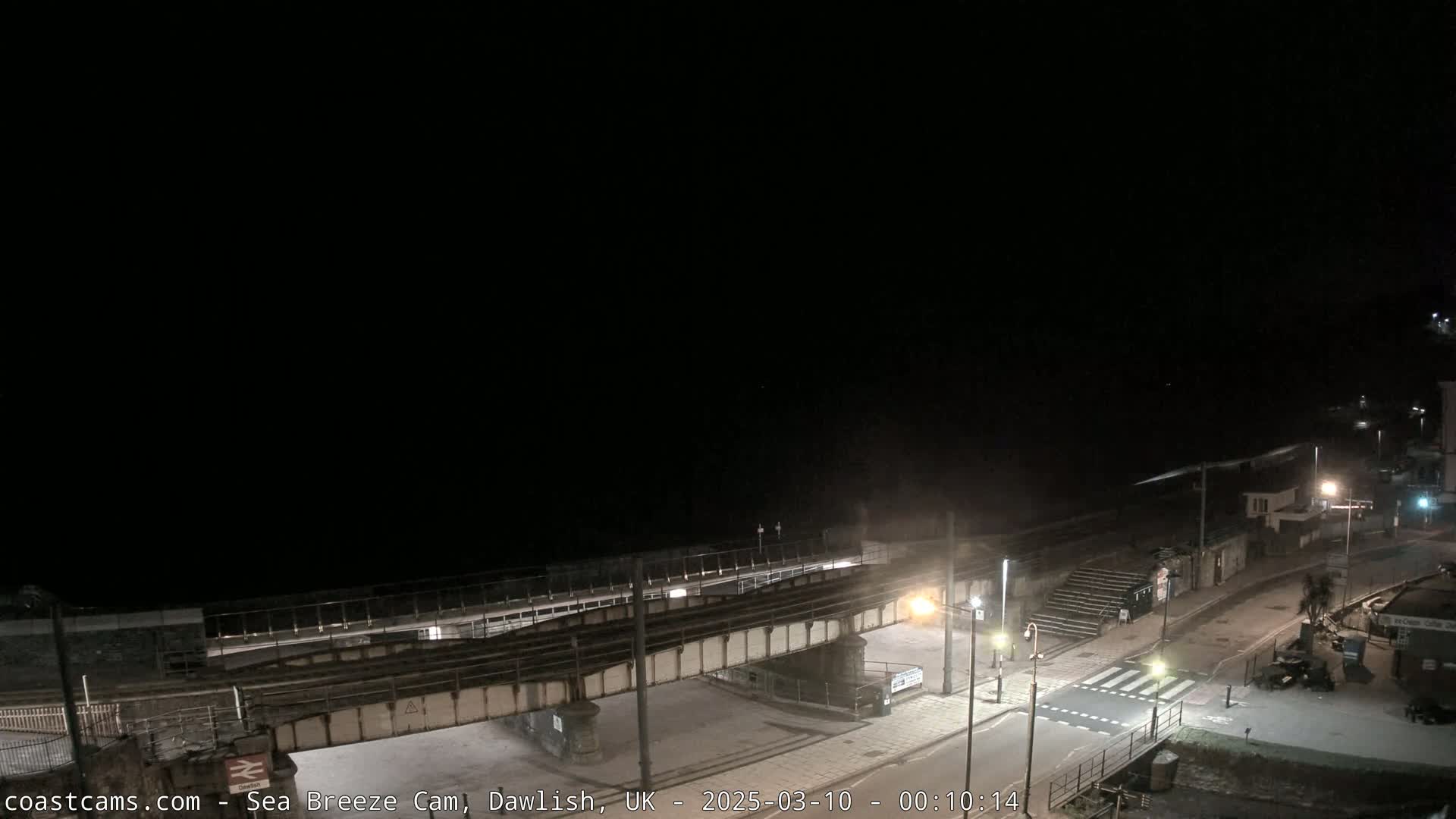 A nighttime view of a coastal road and railway bridge overlooking a dark ocean under a clear, black sky.