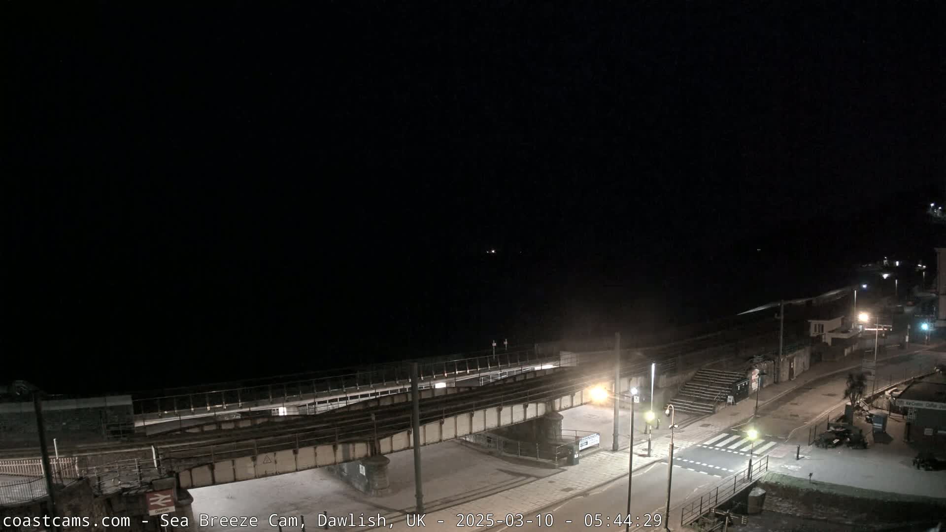 A nighttime, coastal scene shows a dark sea, a street with pedestrian crossing, and a railway bridge over the road.