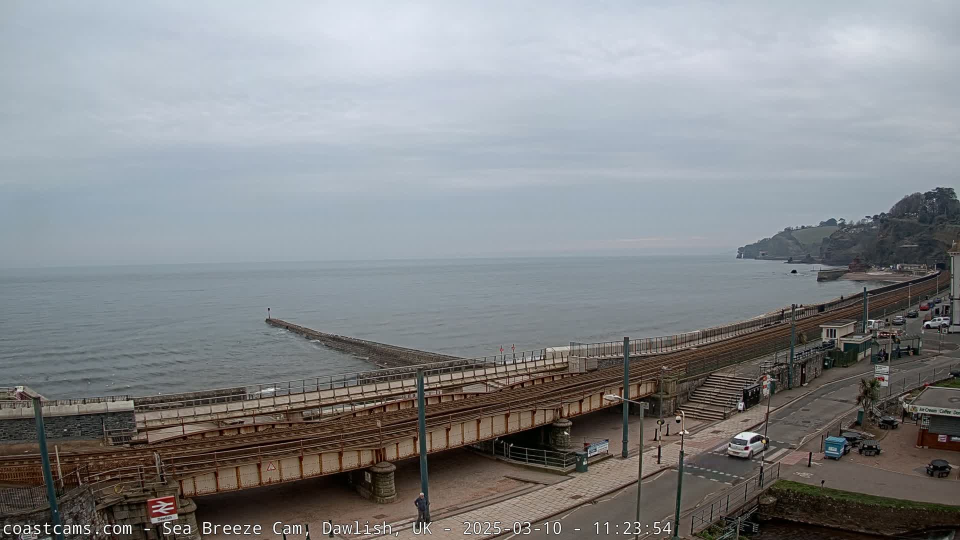A coastal railway line crosses a bridge over a road next to a calm sea under an overcast sky.