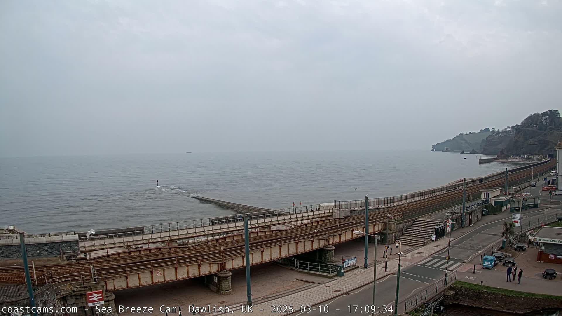 A coastal scene on an overcast day shows a railway line crossing a low bridge over a sea wall and road next to the ocean, with a small town visible on the right.