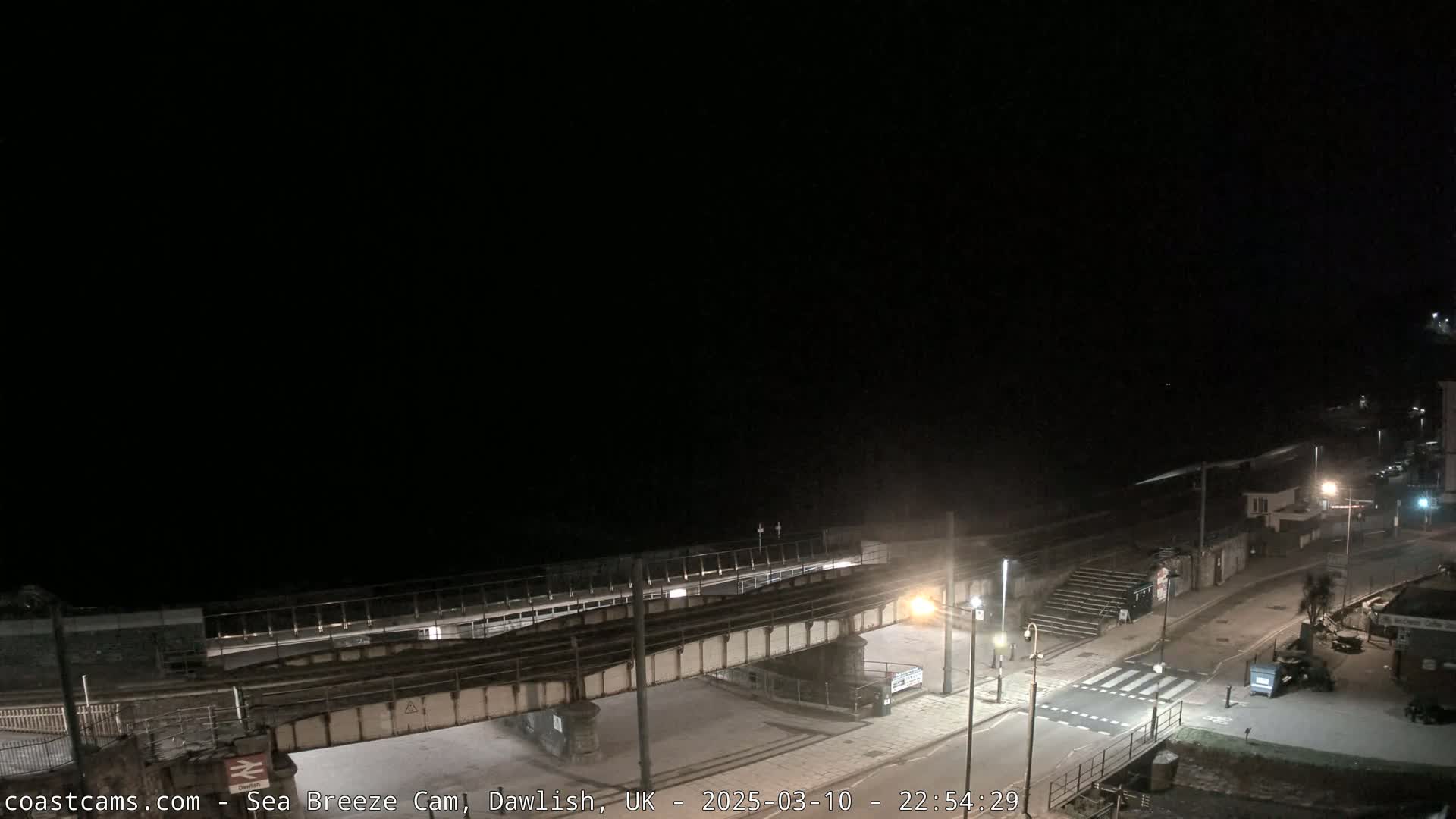 A nighttime view of a coastal town, showing a railway bridge over a road leading to a beach under a dark sky.