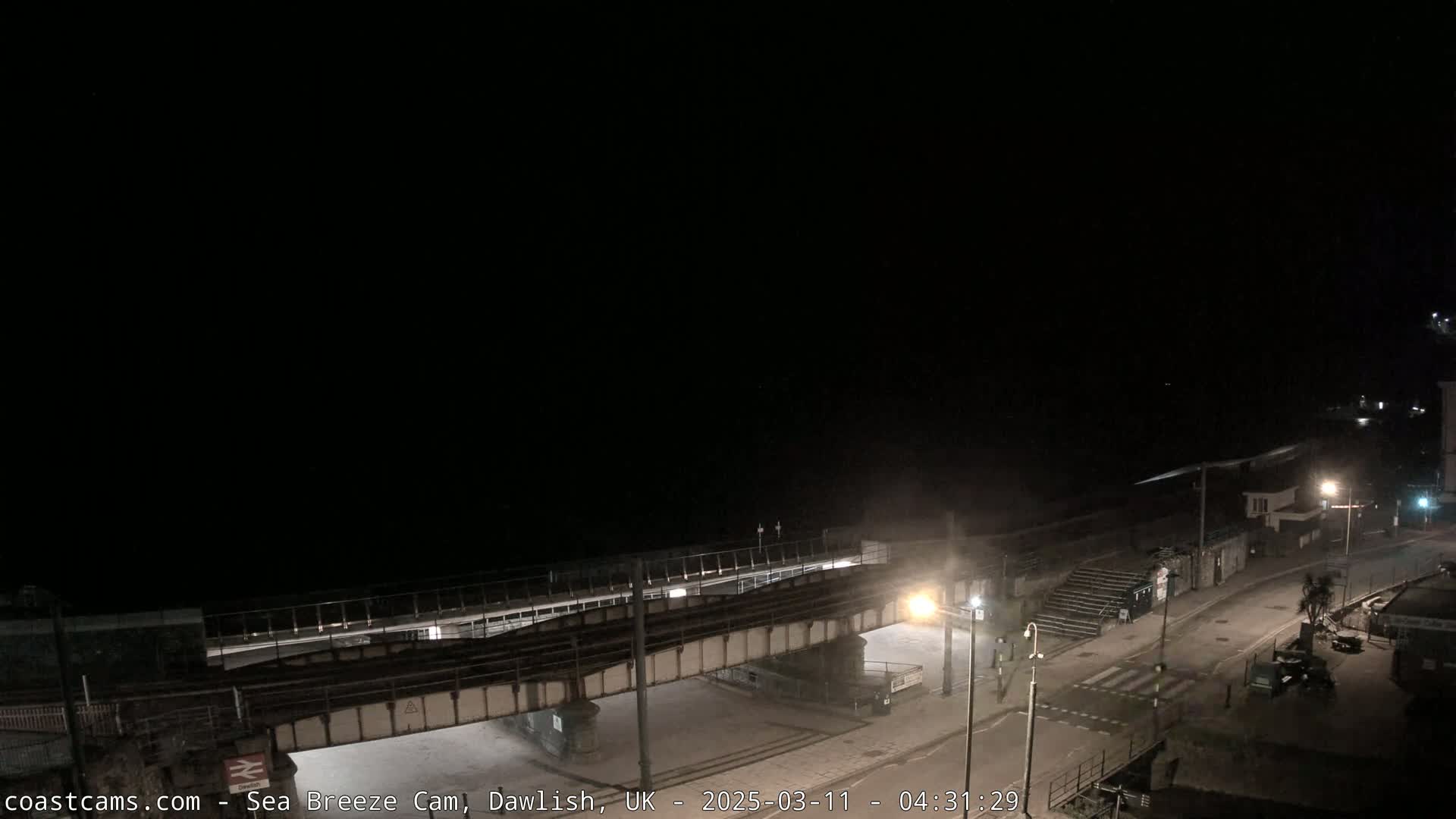 A nighttime view shows a railway bridge over a dark road next to a beach under a clear, dark sky.