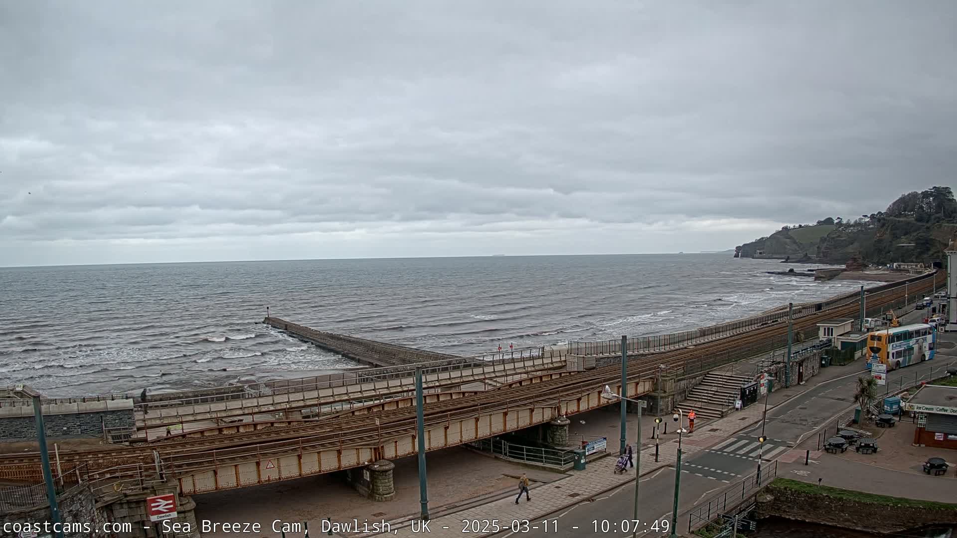 A coastal scene under an overcast sky shows a railway line running along a beach, with a sea wall and small waves breaking on the shore.