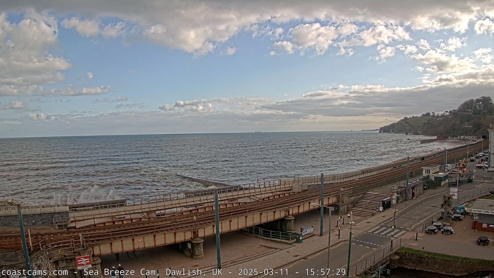 A partly cloudy sky overlooks a choppy sea crashing against a seawall next to railway tracks and a coastal road.
