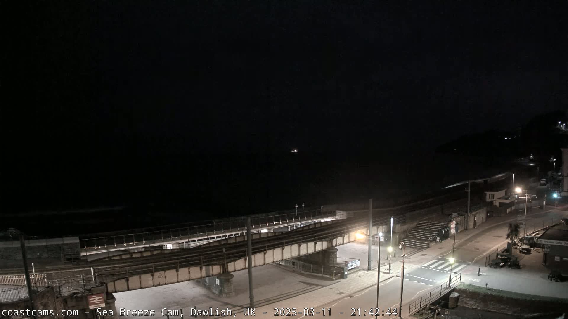 A nighttime coastal scene shows a dark ocean with small waves, a railway bridge over a road leading to a sparsely lit beach, and a few buildings in the distance under a dark sky.