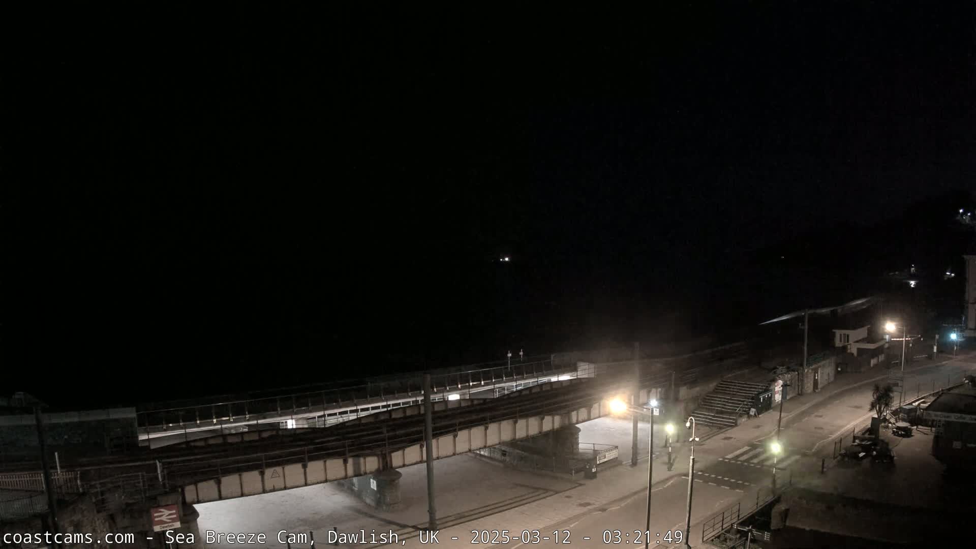 A nighttime, high-angle view shows a train trestle over a road leading to a dark beach and ocean under a clear, black sky.