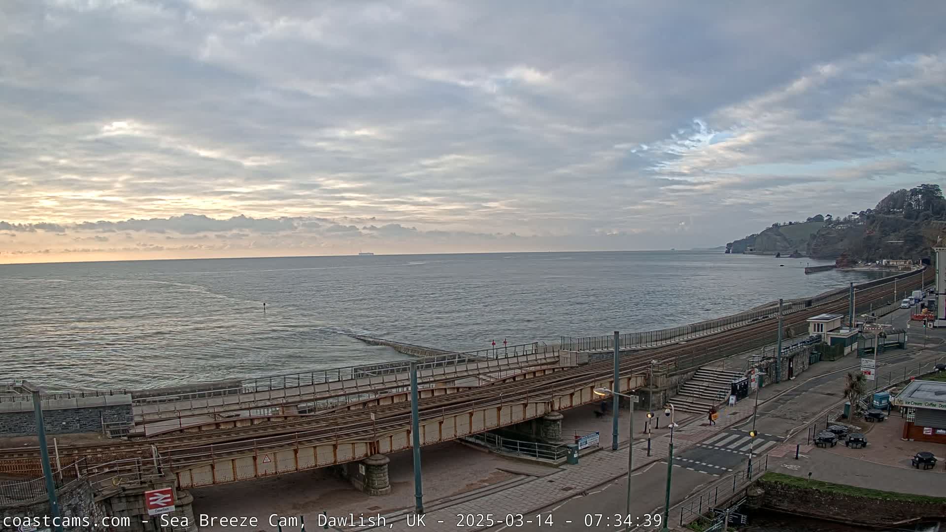 A partly cloudy sunrise over a calm sea, viewed from above a railway bridge crossing a coastal road.