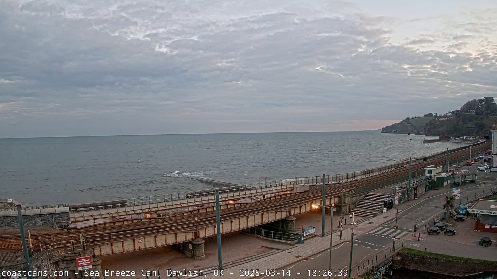 A coastal scene under a cloudy sky shows a train track crossing a seawall next to a road with some buildings and a small beach.
