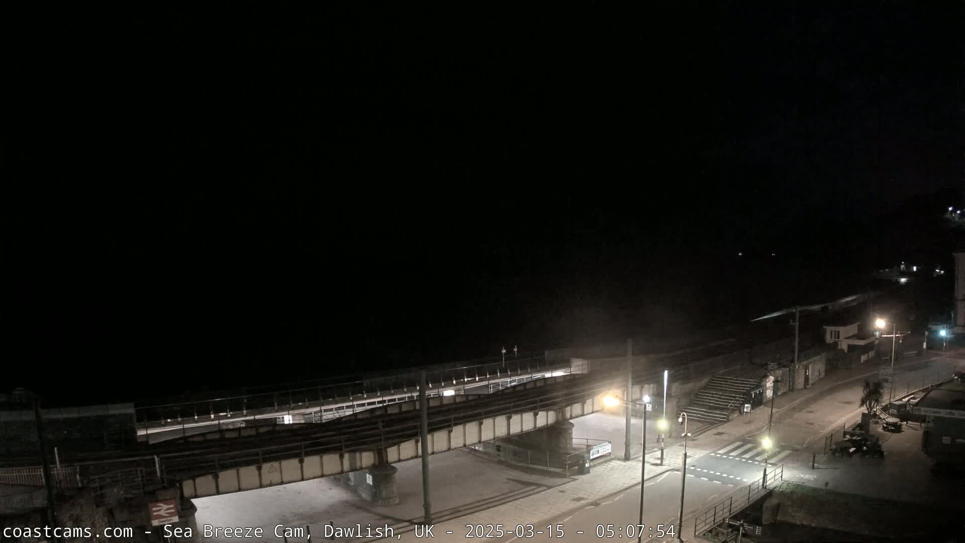 A nighttime view of a coastal town, showing a railway bridge over a road leading to a dark beach under a clear sky.